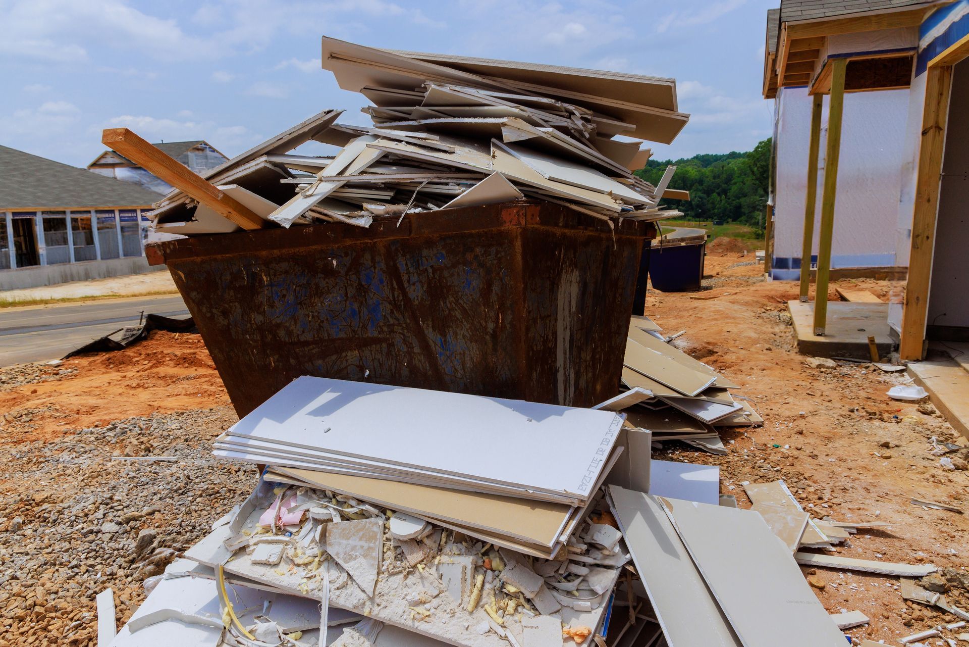 A large, rusted dumpster overflowing with drywall scraps sits on a dirt lot at a construction site.