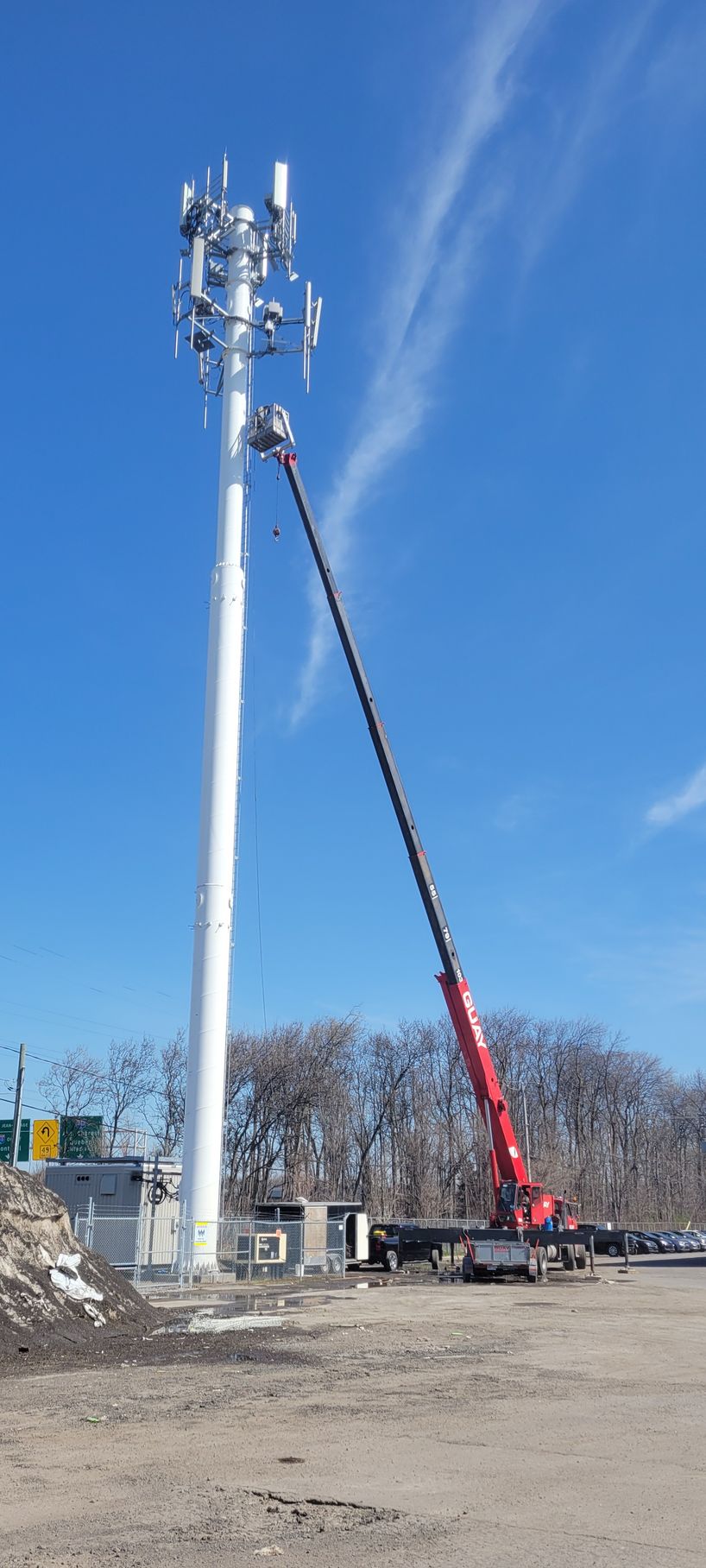 Un camion-grue rouge déploie son bras pour travailler sur une haute tour de télécommunications cellulaire blanche se détachant sur un ciel bleu clair.