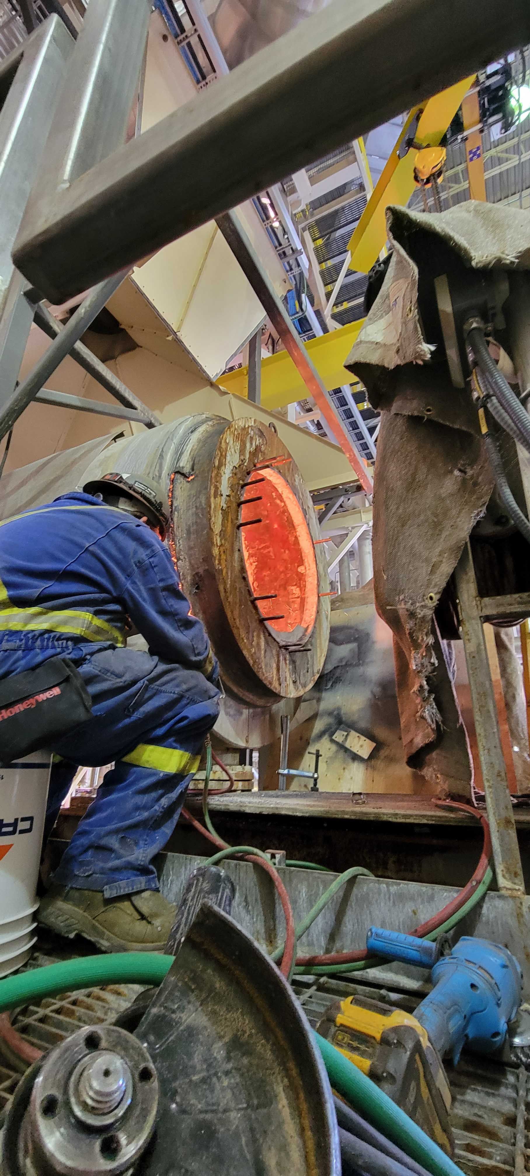 Dans un atelier, un ouvrier en tenue de protection est accroupi près d'un four industriel incandescent.