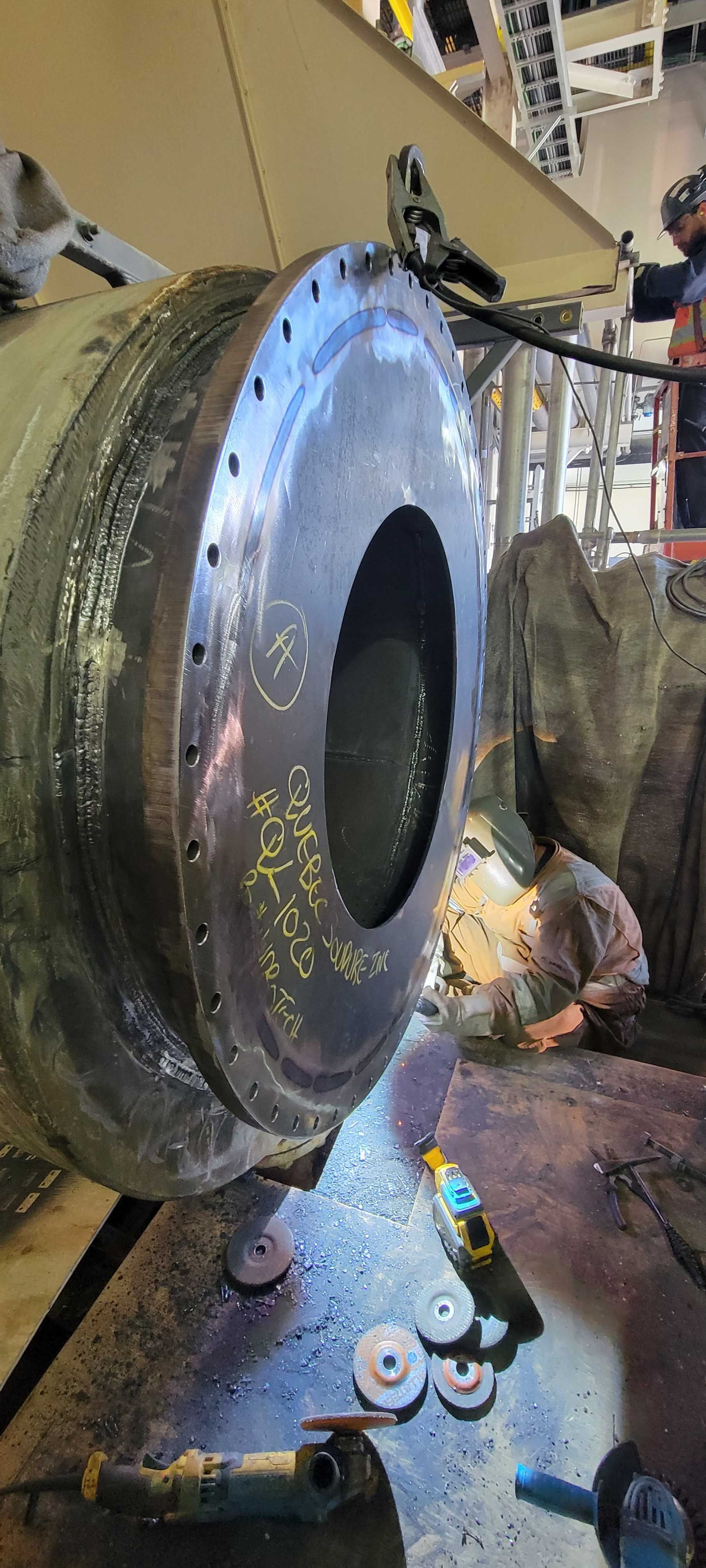 Dans un atelier industriel, une personne portant un équipement de protection soude une grande bride métallique, ses outils étant éparpillés au sol.