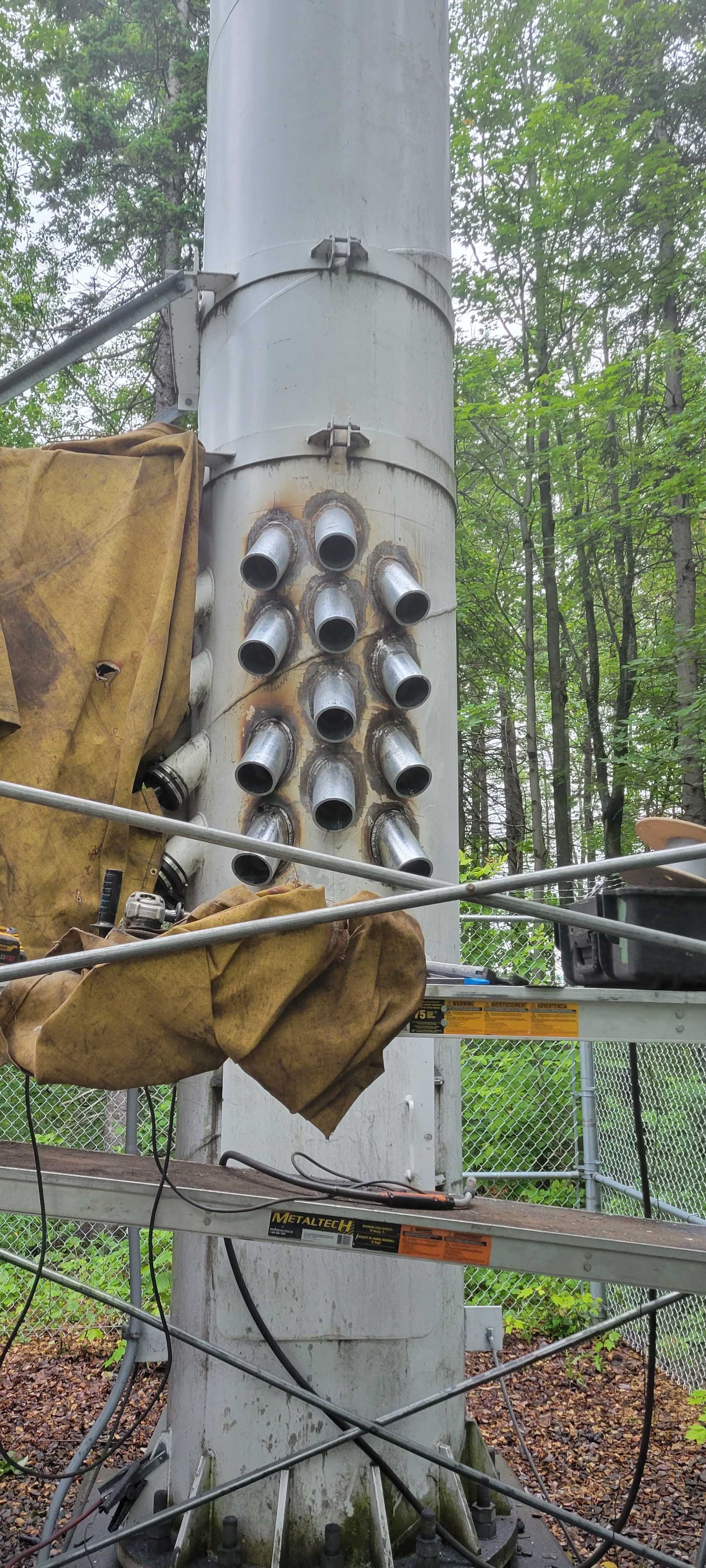 Un poteau électrique vertical blanc est en cours de réparation à l'extérieur, avec une série de rallonges cylindriques métalliques soudées.