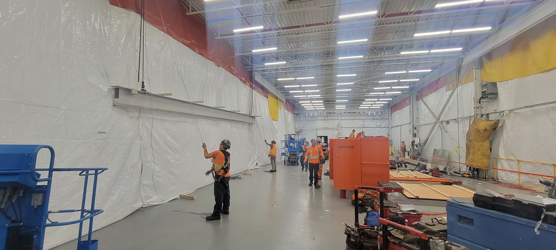 Des ouvriers vêtus de tenues orange haute visibilité assemblent ou inspectent un long rail métallique dans une grande installation industrielle intérieure.