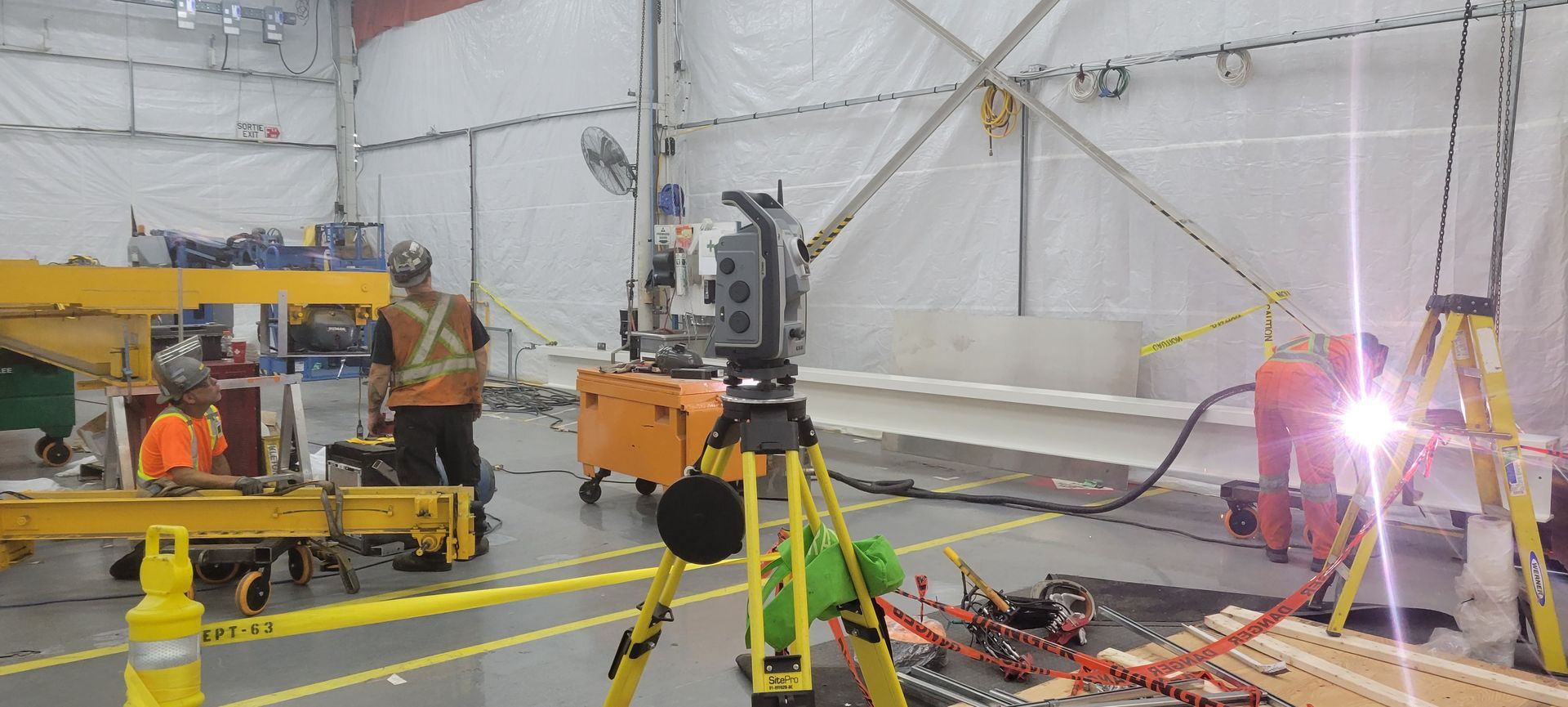 Des ouvriers du bâtiment utilisent une station totale de levé topographique dans un grand espace industriel recouvert d'une tente blanche, où des travaux de soudure sont en cours.