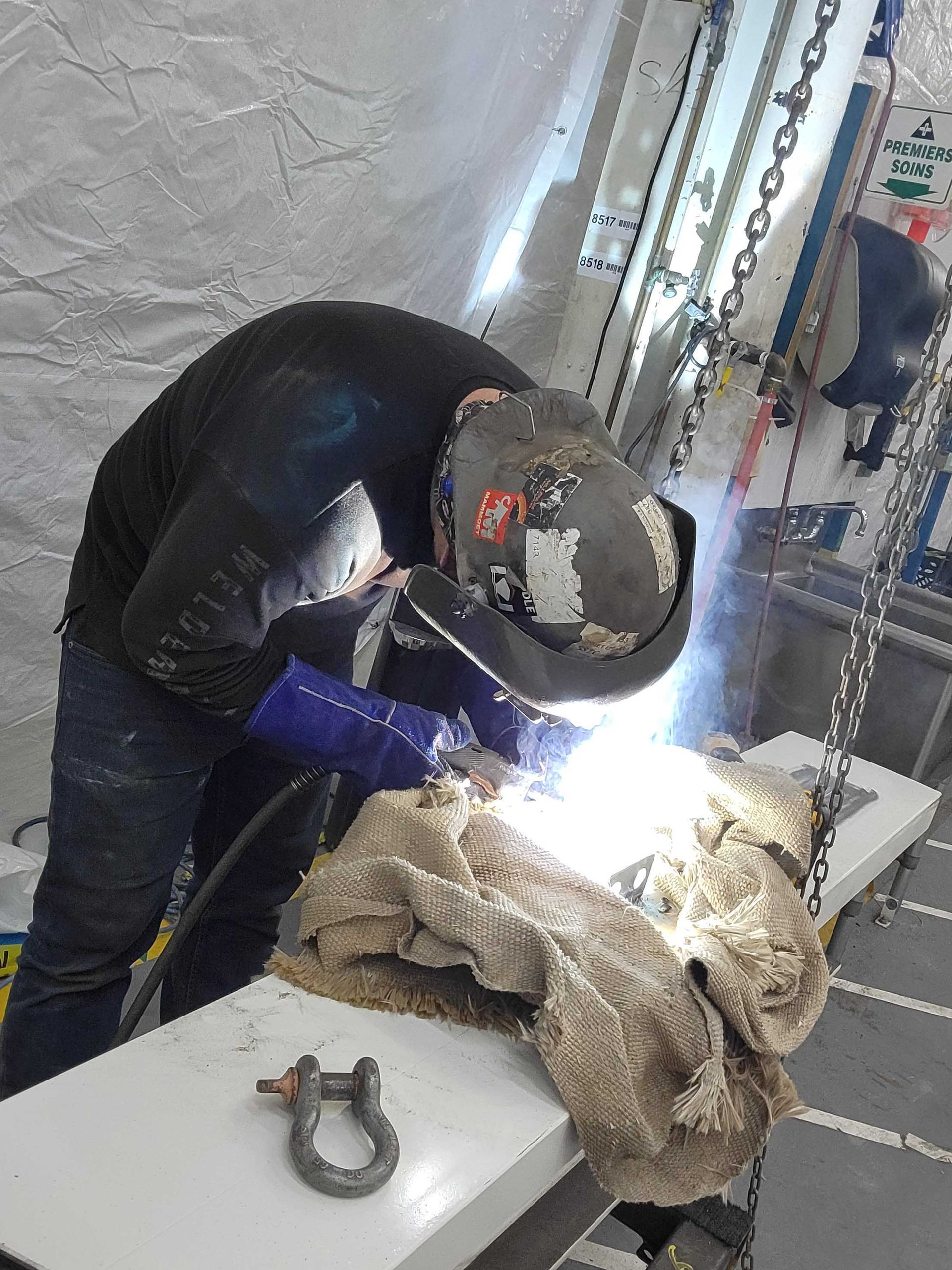 Une personne en tenue de protection soude des pièces métalliques dans un atelier, la lumière vive de la torche éclairant le lieu.