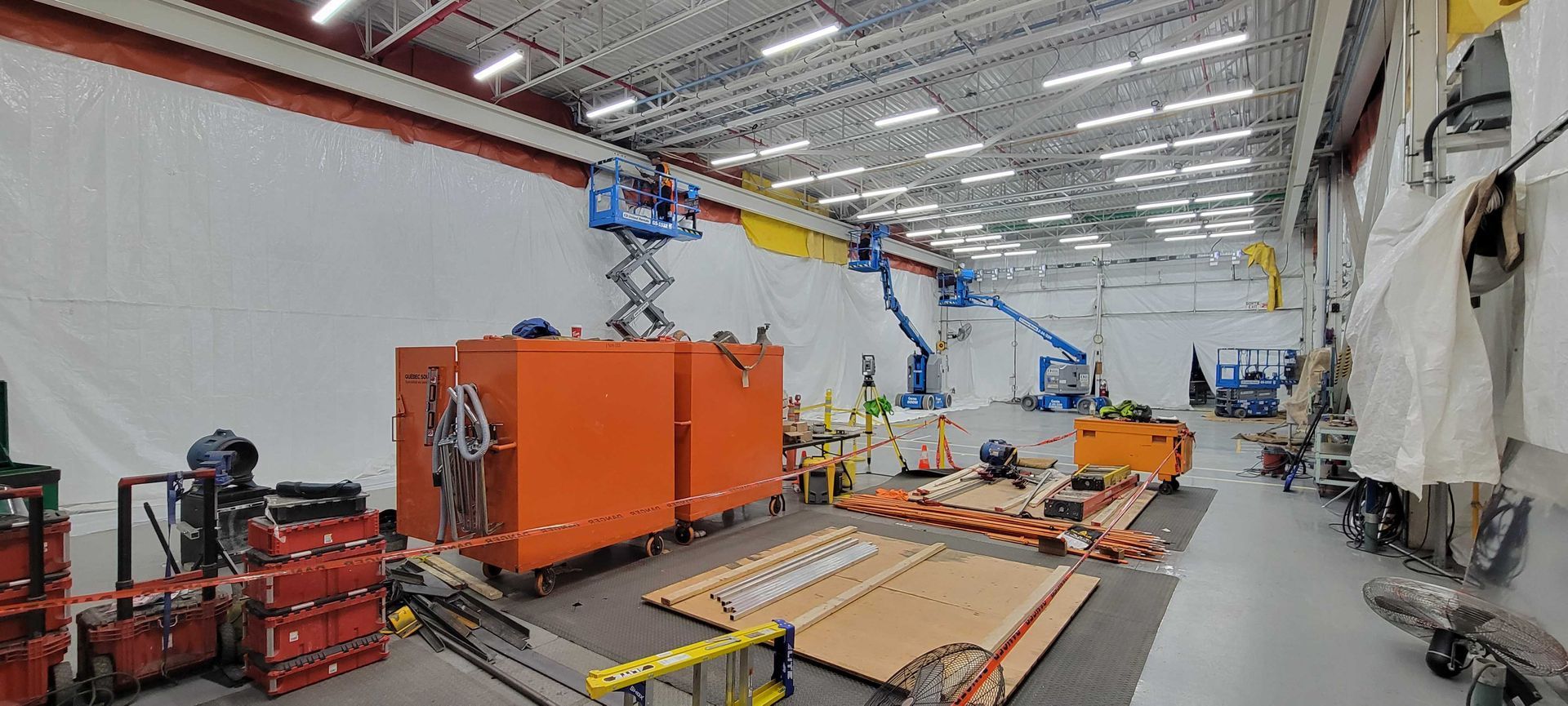 Vue grand angle d'un vaste entrepôt industriel avec des outils de construction, des conteneurs de stockage orange et des nacelles élévatrices.