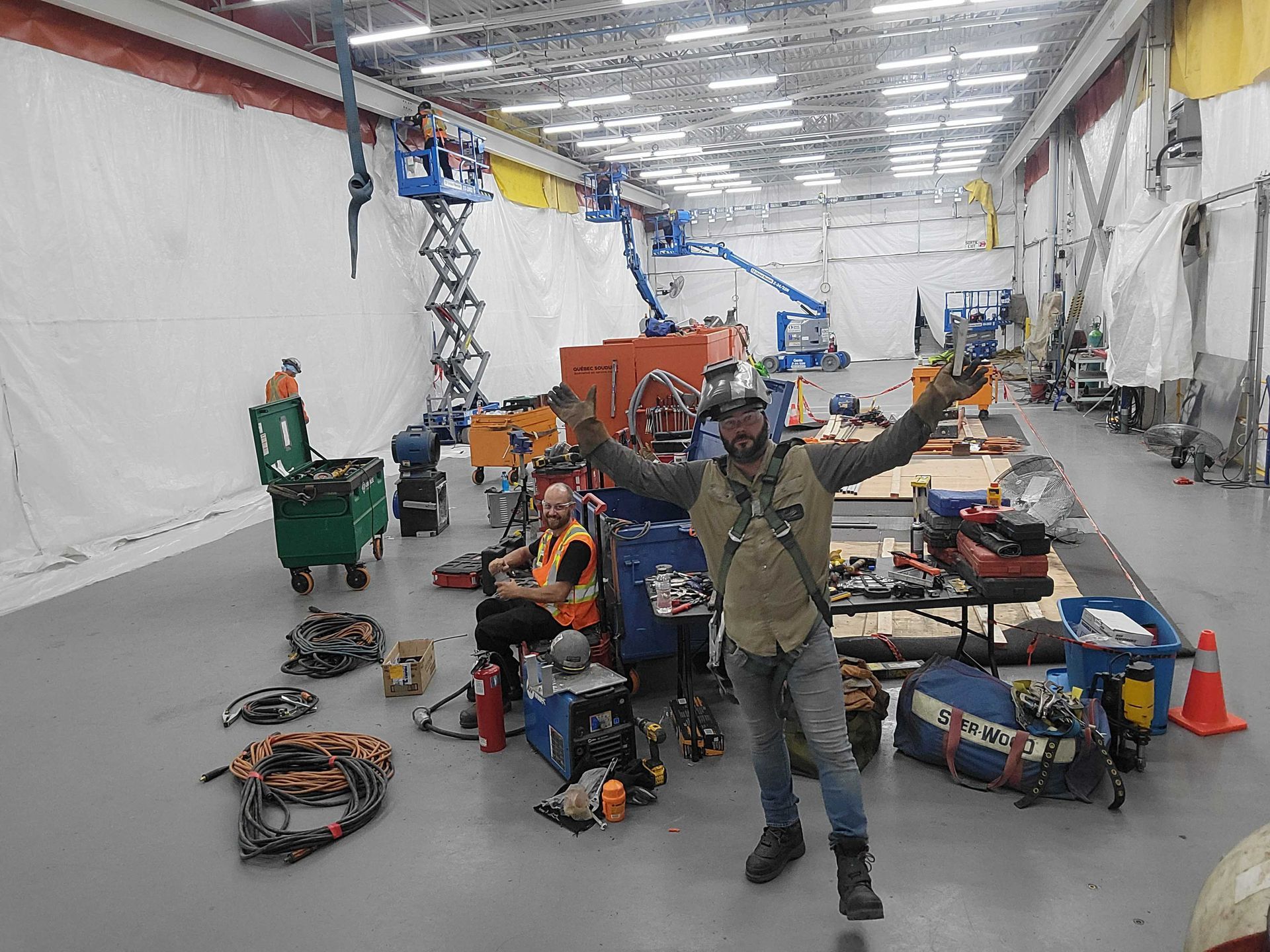 Un ouvrier en tenue de sécurité se tient debout, les bras levés, dans un grand atelier industriel où l'on aperçoit des outils et des ponts élévateurs en arrière-plan.
