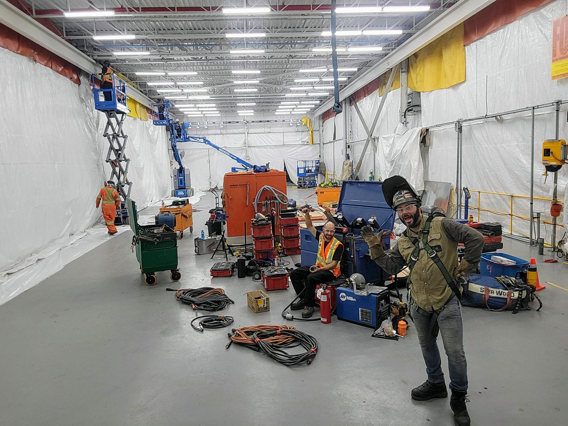 Des ouvriers en tenue de sécurité et masque de soudeur se tiennent debout dans un grand espace industriel où se trouvent des équipements lourds et des murs blancs drapés.