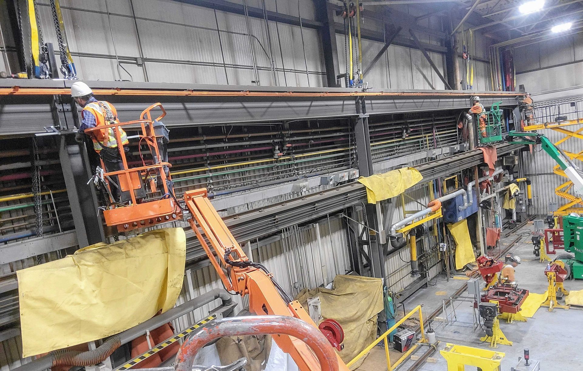 Des ouvriers portant des gilets haute visibilité utilisent des nacelles élévatrices pour installer ou entretenir des câbles sur une grande structure industrielle.