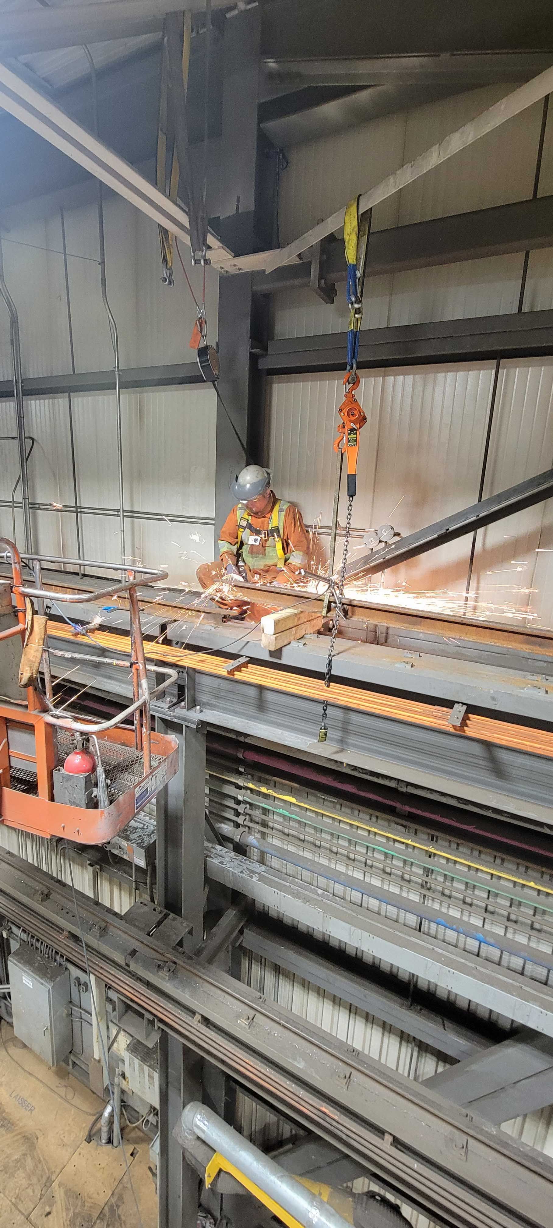 Un ouvrier portant un gilet haute visibilité et un harnais de sécurité effectue des travaux de maintenance à proximité d'équipements industriels suspendus.