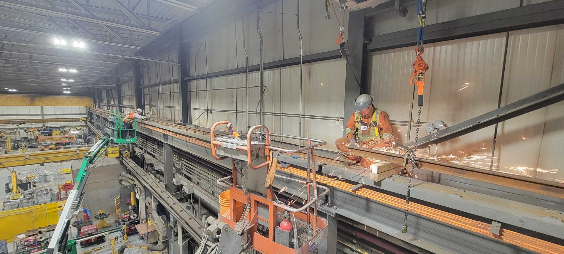 Un ouvrier portant un harnais de sécurité effectue des travaux à chaud sur des poutres d'acier en hauteur à l'intérieur d'un grand entrepôt industriel éclairé.