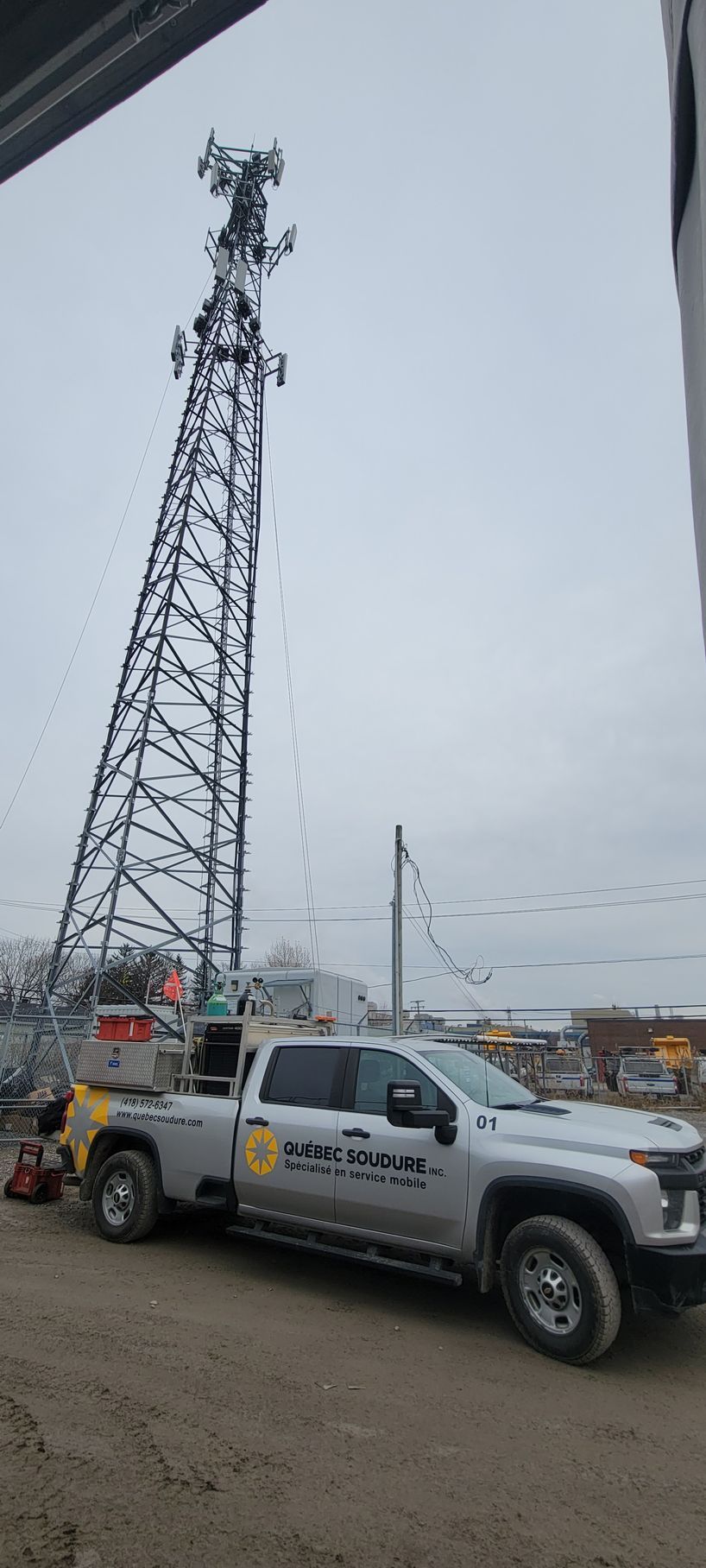 Un camion utilitaire argenté est garé sur un terrain vague à côté d'une haute tour de télécommunications, sous un ciel couvert.