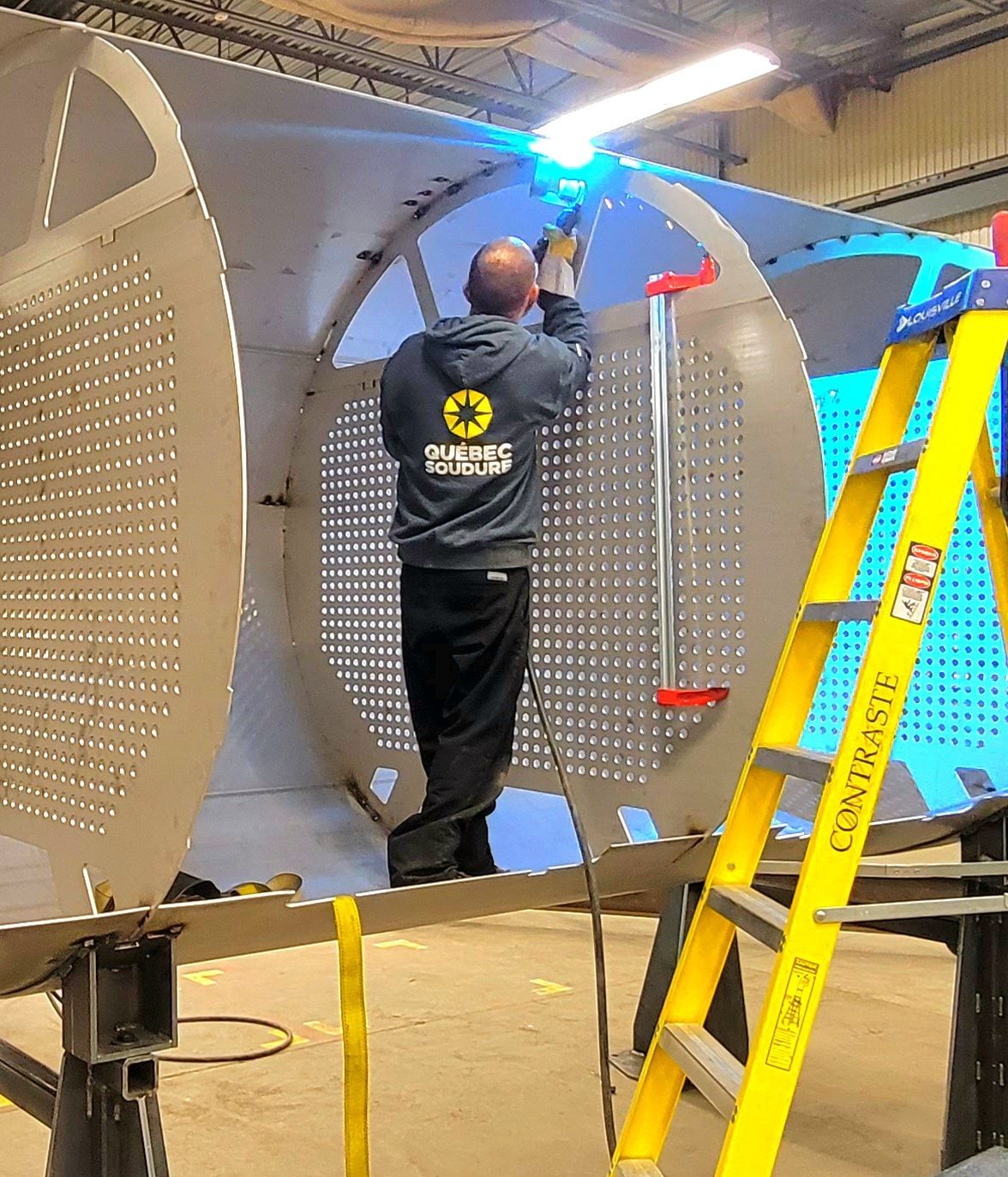 Une personne portant un sweat à capuche gris soude une structure métallique à panneau perforé à l'intérieur d'un atelier.