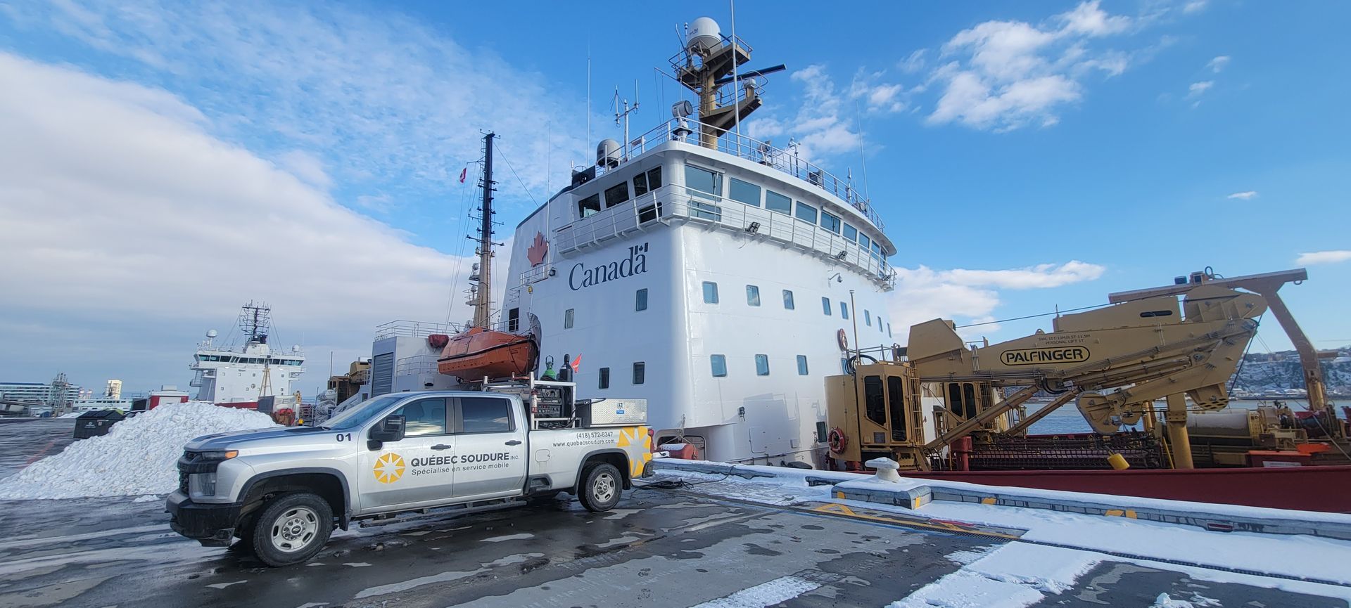 Une camionnette blanche garée sur un quai enneigé, à côté d'un grand brise-glace blanc, sous un ciel d'un bleu éclatant.