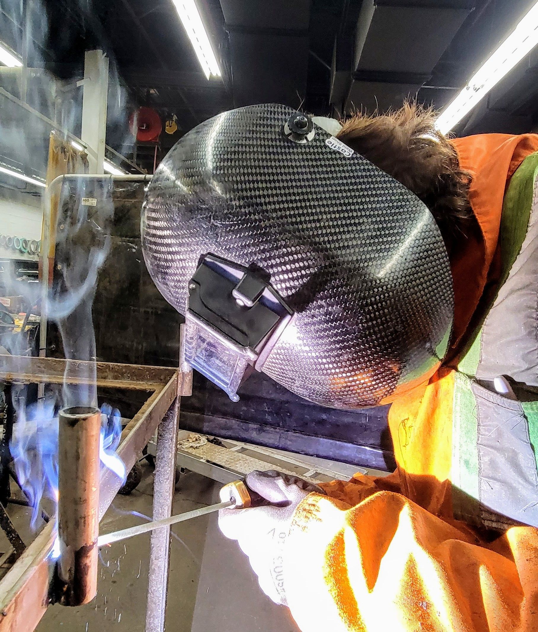 Une personne portant un casque de soudeur réfléchissant et un équipement haute visibilité se concentre sur la soudure d'un tuyau métallique dans un atelier.