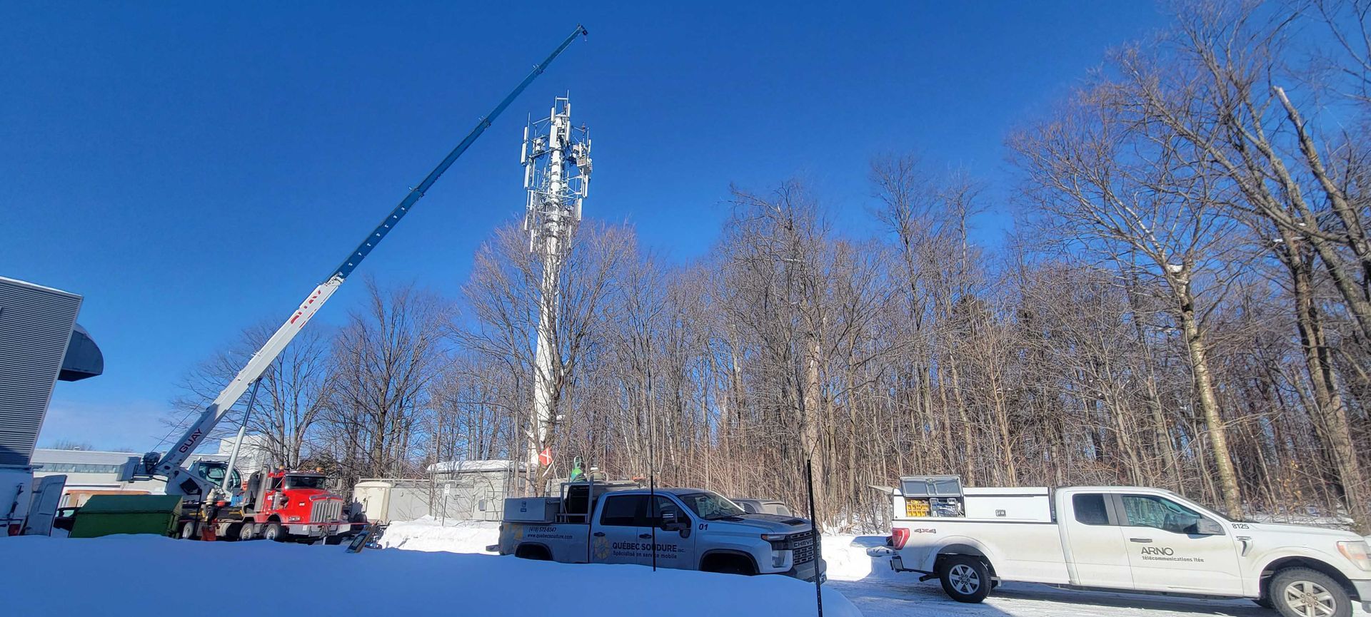 Un camion-grue et des véhicules utilitaires travaillent sur un site de pylône de téléphonie mobile entouré d'un sol enneigé et d'arbres dénudés.