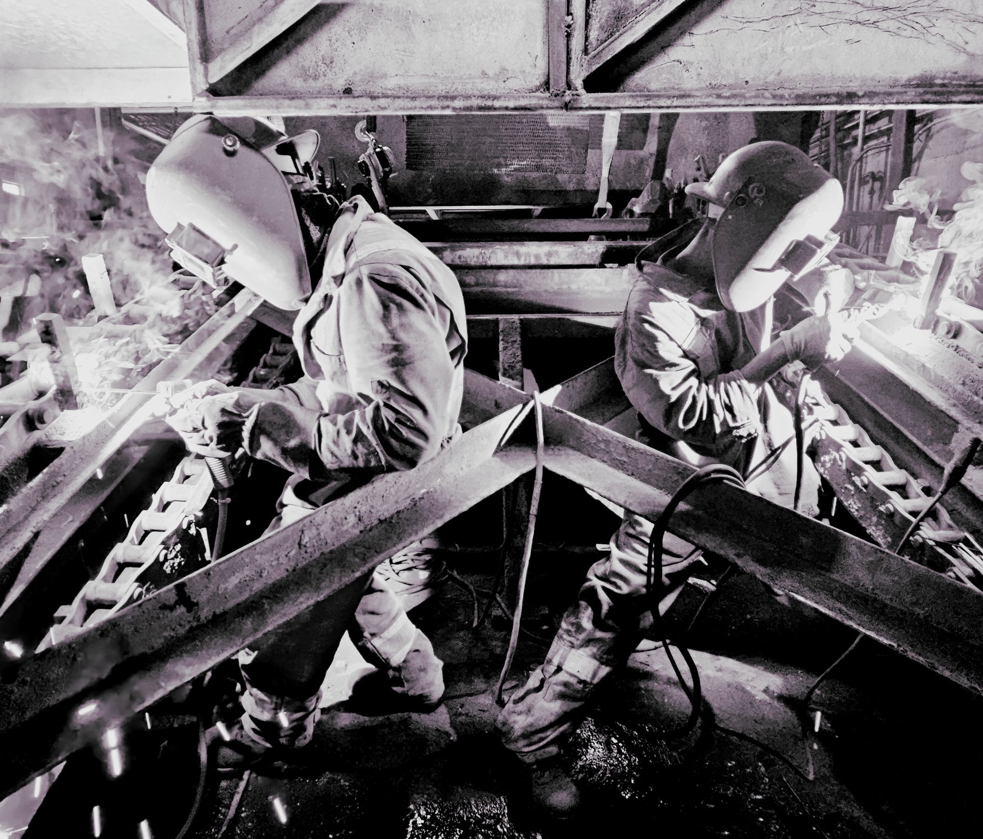 Deux soudeurs portant des casques de protection travaillent sur une structure métallique dans un environnement industriel faiblement éclairé.