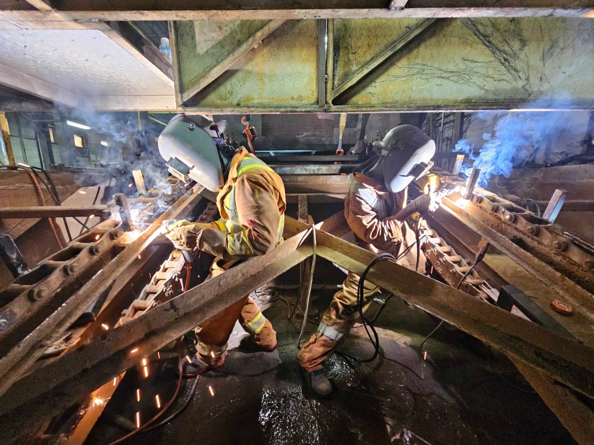 Deux soudeurs, vêtus de tenues de protection et de casques, travaillent sur des poutres métalliques dans un environnement industriel, produisant des étincelles brillantes.