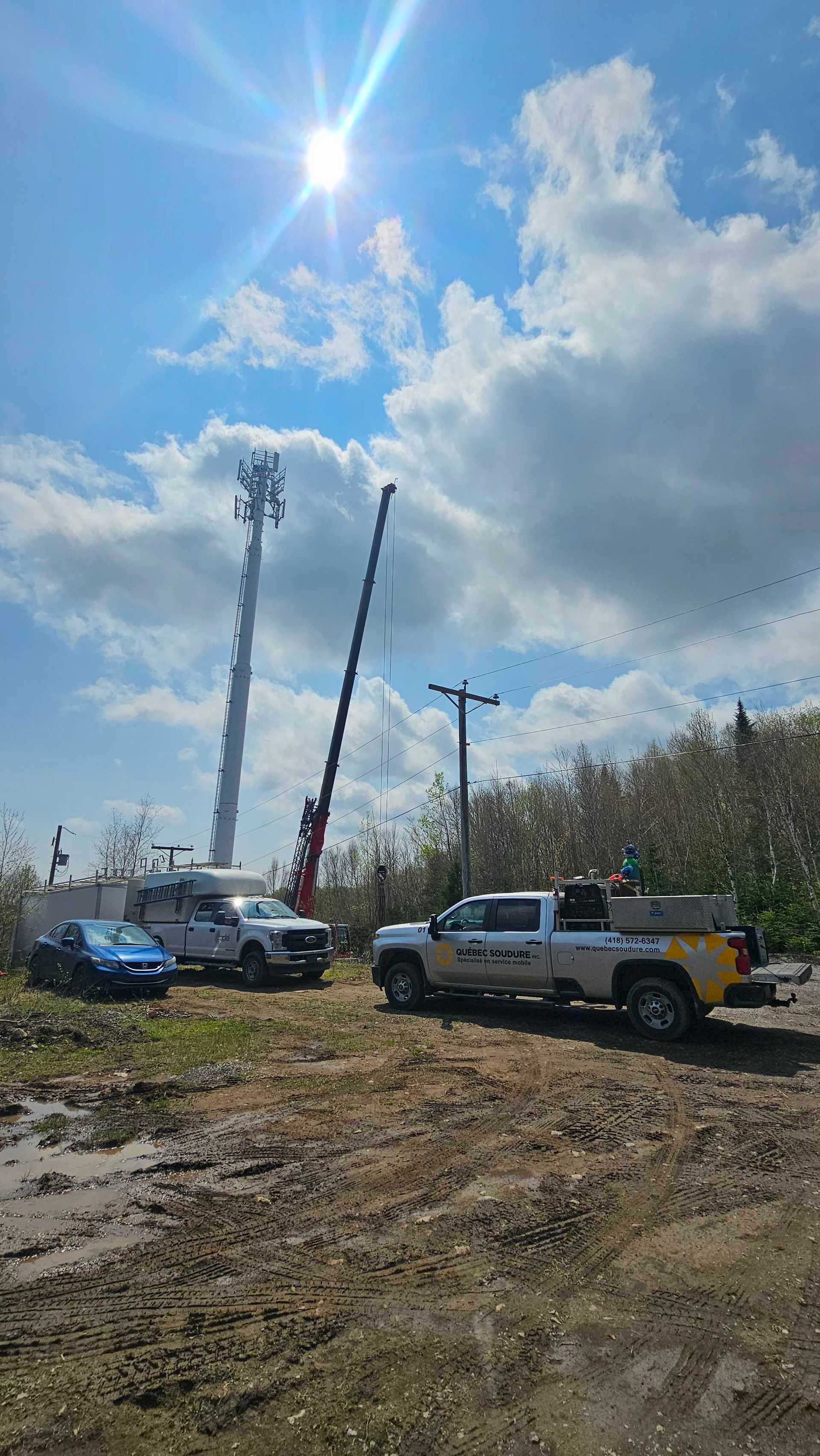Un chantier de construction avec deux camions et une grue stationnés près d'une haute tour de télécommunications sous un ciel ensoleillé et nuageux.