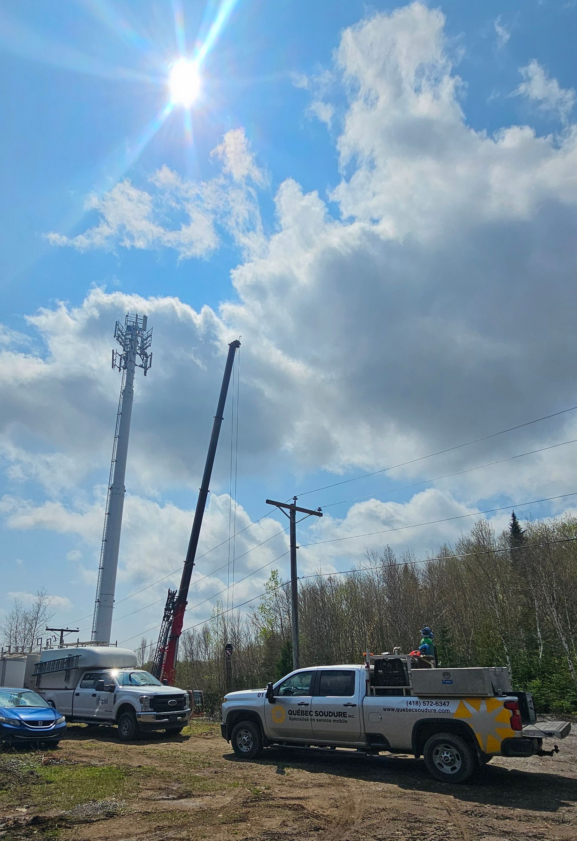 Une grue travaille sur un haut pylône de téléphonie mobile sous un soleil éclatant, avec deux camions utilitaires stationnés au premier plan.
