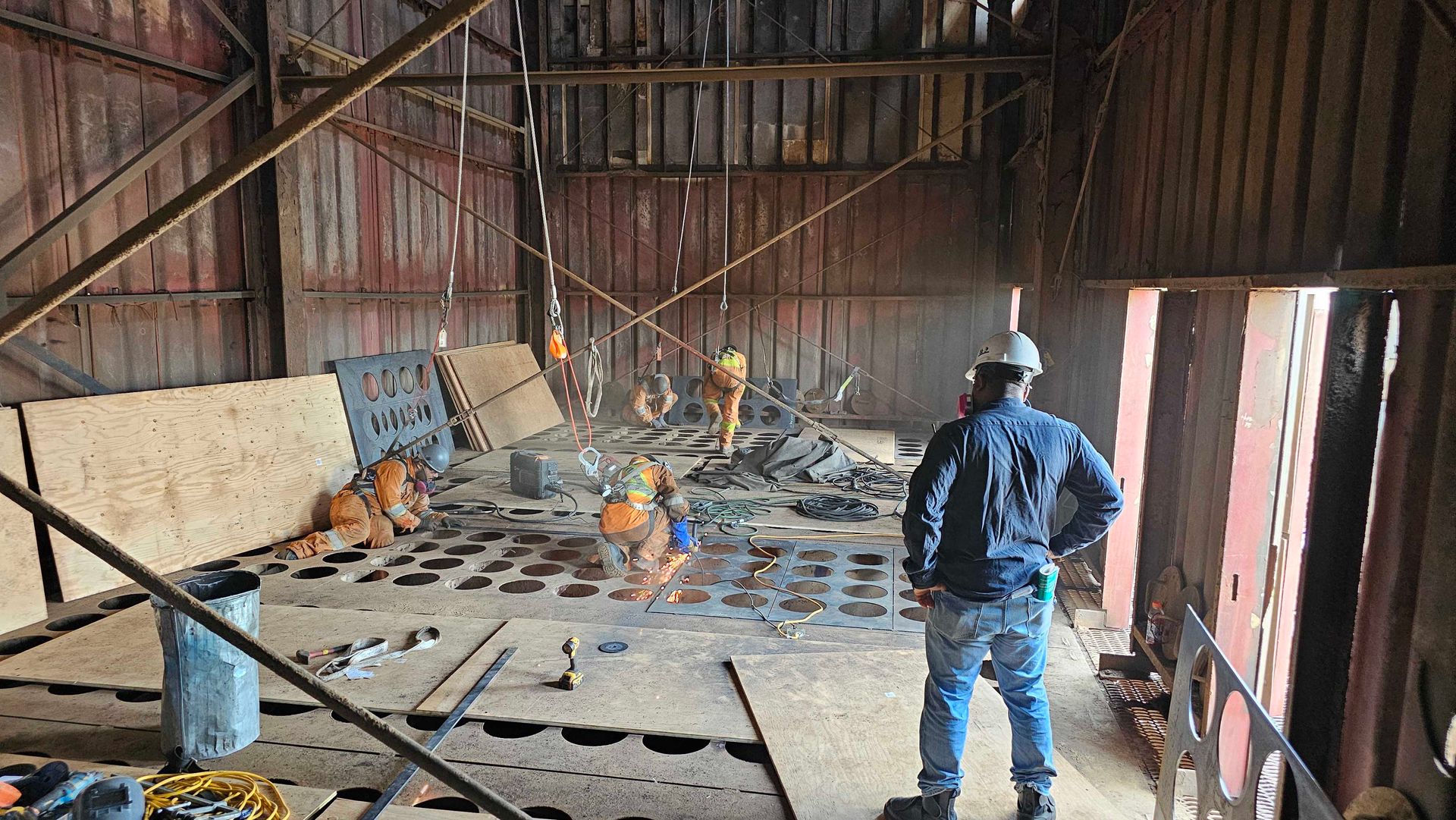 Un ouvrier du bâtiment observe une équipe en train de souder et d'assembler des composants métalliques à l'intérieur d'une structure industrielle inachevée.