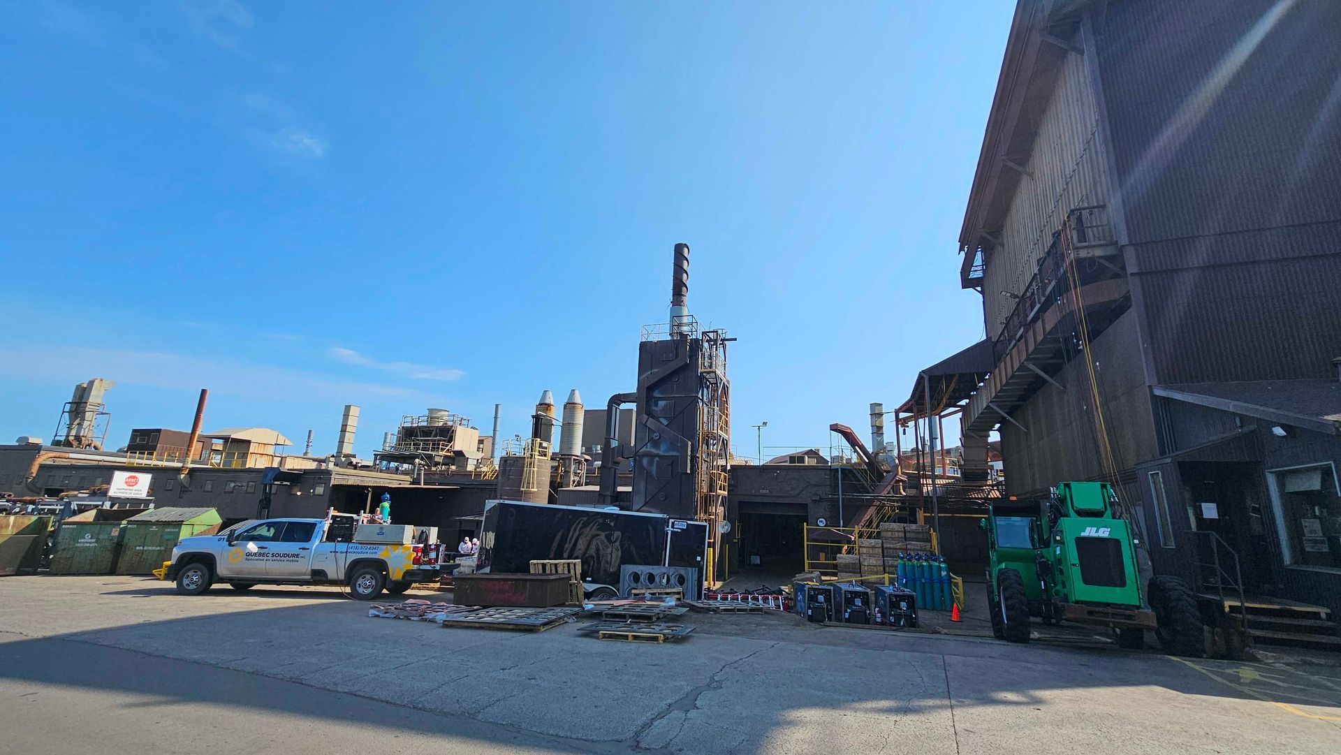 Vue grand angle d'un bâtiment industriel partiellement démoli et délabré, sous un ciel bleu dégagé, avec des véhicules stationnés.