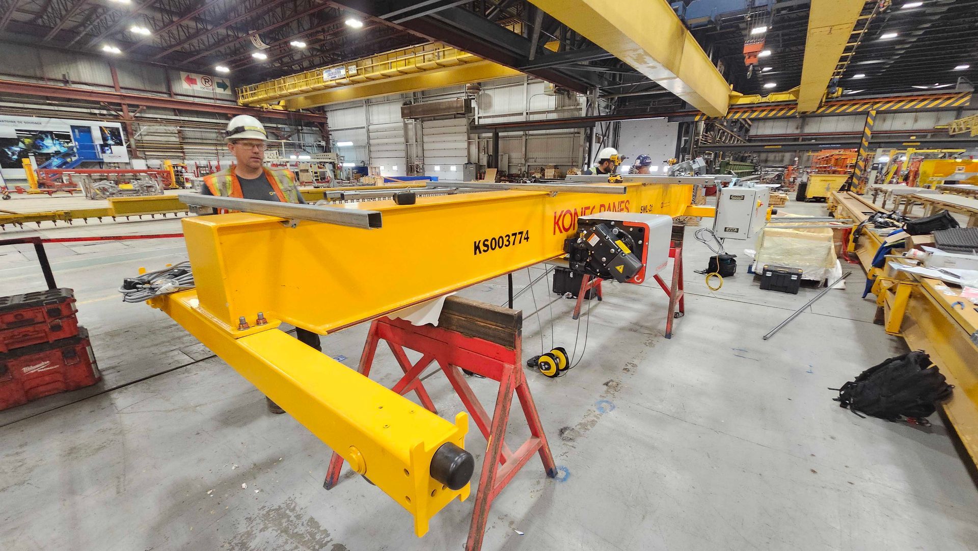 Un ouvrier inspecte dans un entrepôt une grande poutre jaune de grue industrielle reposant sur des tréteaux métalliques rouges.