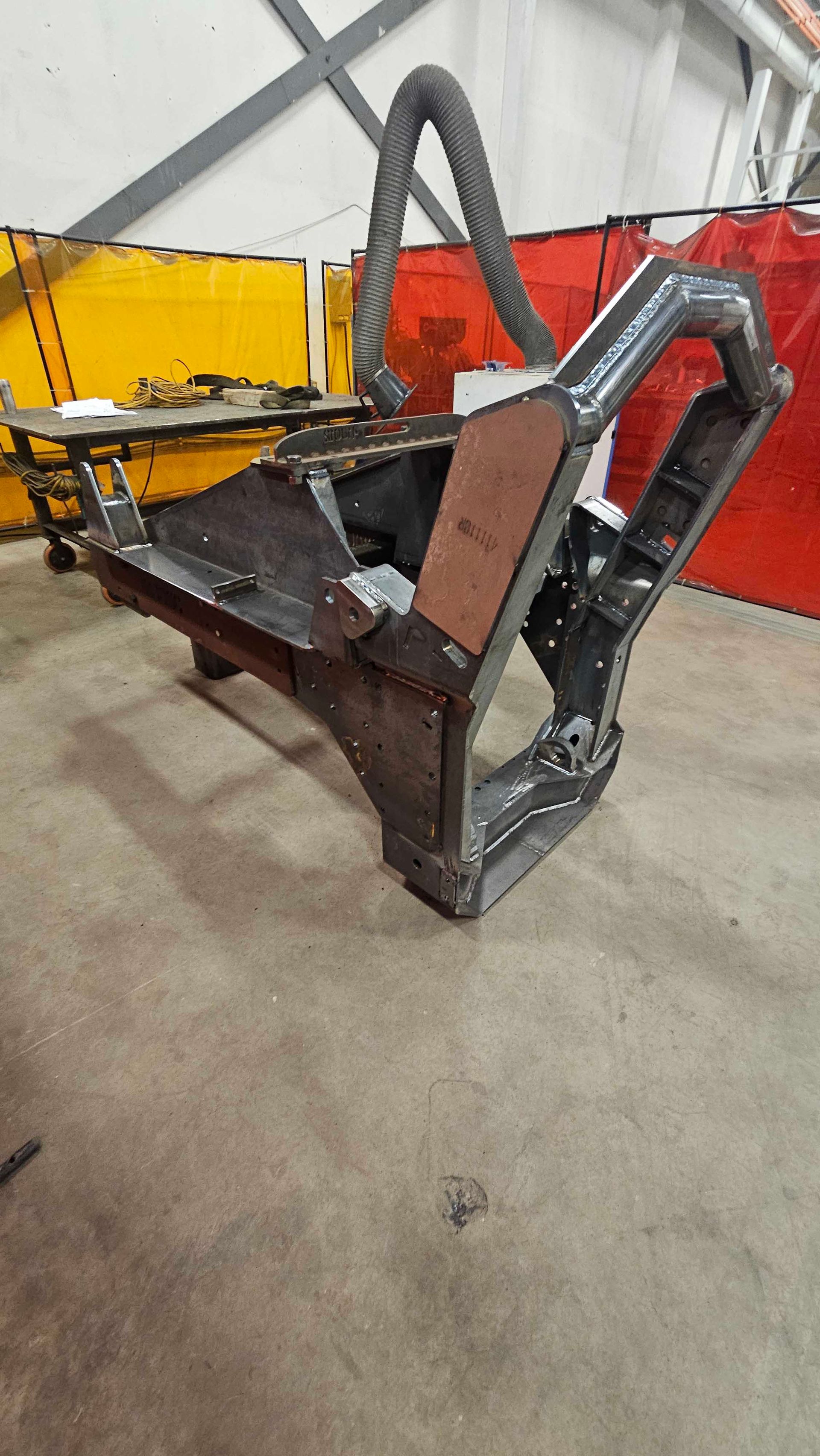 Dans un atelier aux vitres de sécurité colorées, une imposante structure métallique sombre, semblable à celle d'une machine industrielle, se dresse sur un sol en béton.
