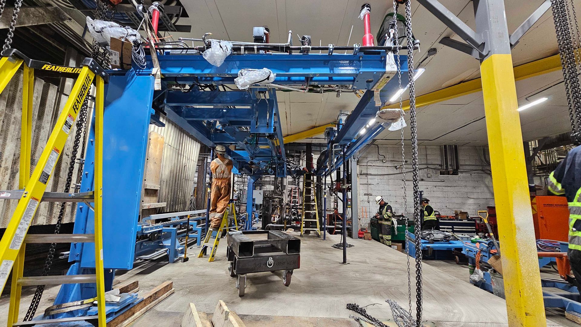 Entrepôt industriel où des ouvriers installent une grande structure de pont roulant bleu au-dessus d'un sol en béton.
