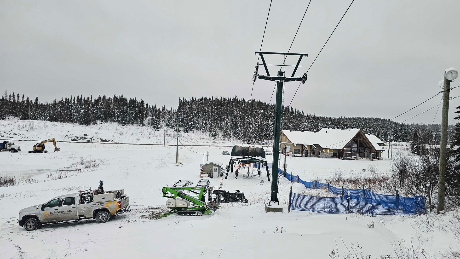 Au pied des remontées mécaniques, recouvert de neige, on aperçoit un camion d'entretien, un véhicule vert servant à la maintenance des remontées et, au loin, un chalet.