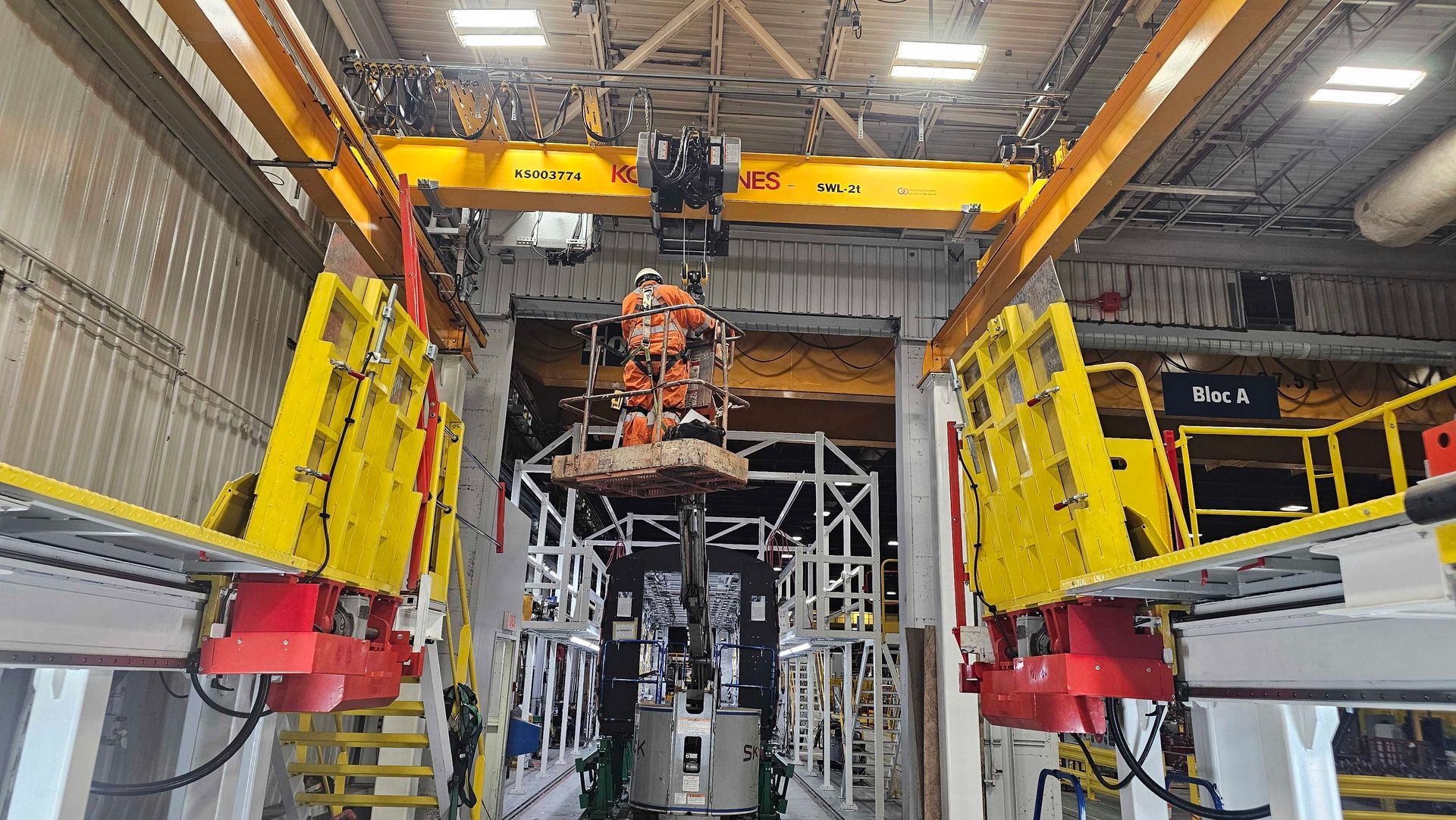 Une personne portant un équipement de sécurité est suspendue par un pont roulant jaune à l'intérieur d'une usine de fabrication industrielle.