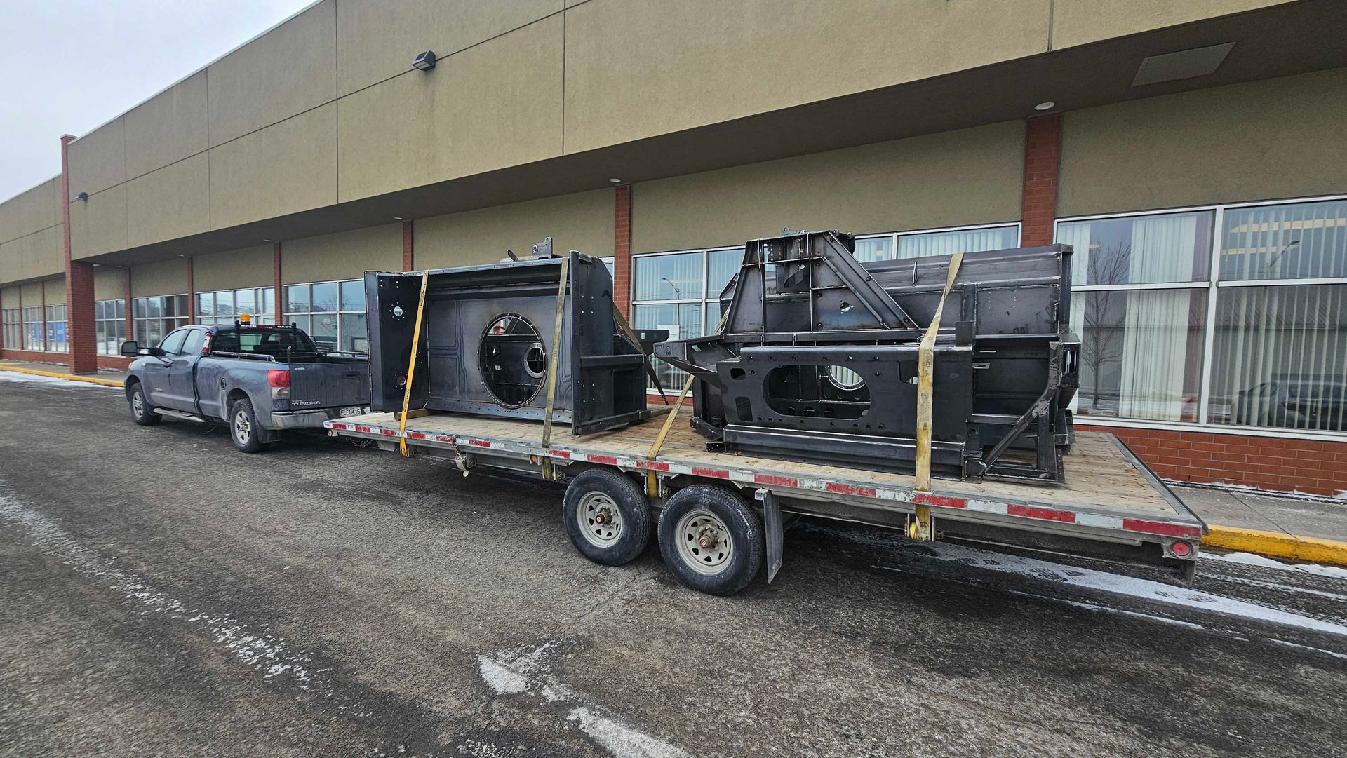 Une camionnette grise tracte une remorque à plateau chargée de deux gros composants métalliques industriels noirs devant un bâtiment.
