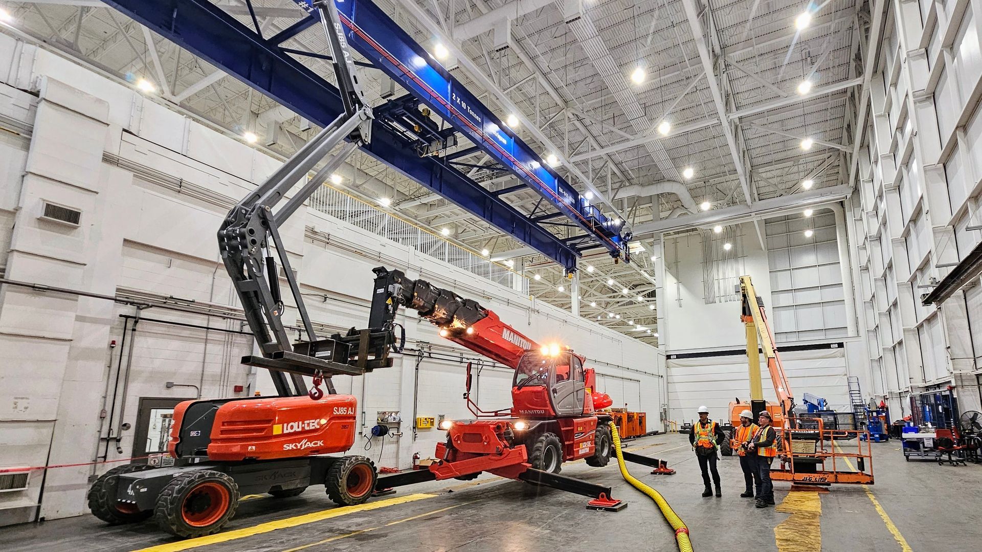 Deux plateformes de travail aériennes rouges et du personnel travaillant à l'intérieur d'un grand entrepôt industriel avec des poutres de pont roulant.