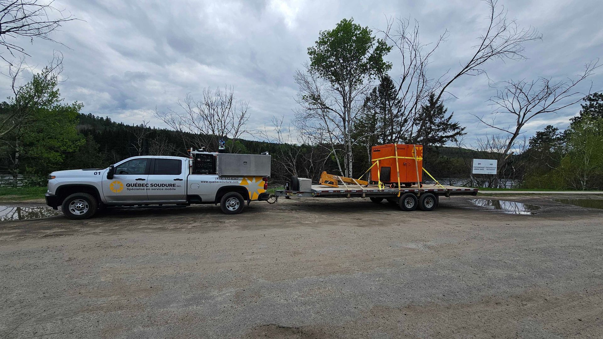 Une camionnette argentée à benne utilitaire tire une remorque à plateau transportant un conteneur orange sur un terrain gravillonné.