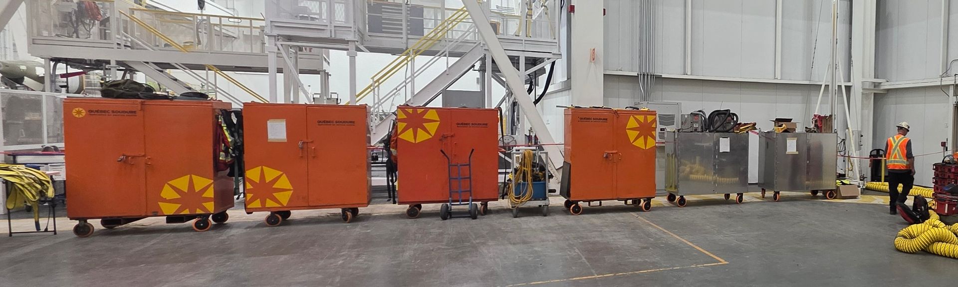 Une rangée de conteneurs industriels orange ornés d'un logo en forme de fleur jaune, sur un sol en béton dans une installation industrielle.
