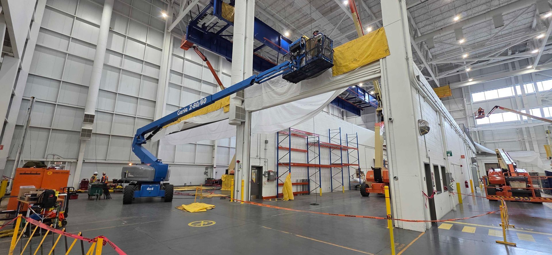 Dans un grand entrepôt industriel, une nacelle élévatrice bleue s'élève vers le ciel, où des ouvriers l'utilisent pour installer une poutre aérienne jaune.