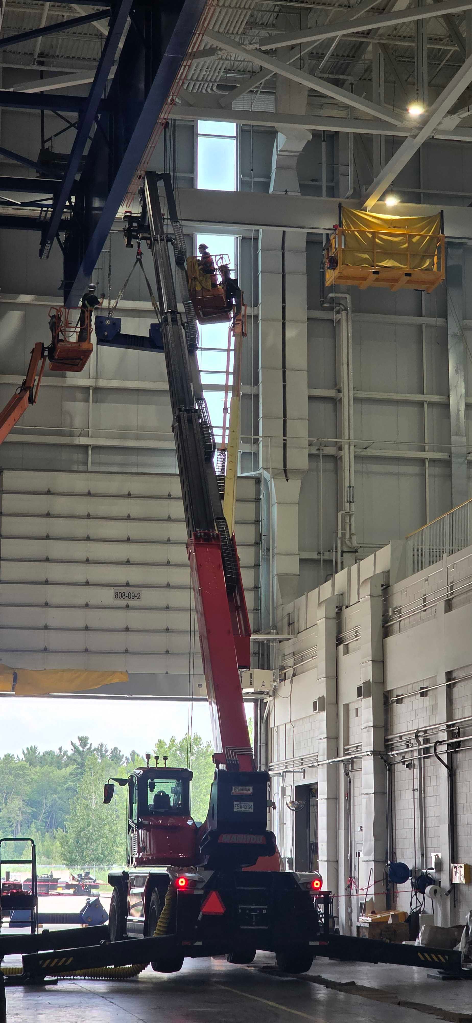 Une grue mobile rouge à flèche déployée se trouve à l'intérieur d'un bâtiment industriel, soulevant un conteneur jaune vers le plafond.