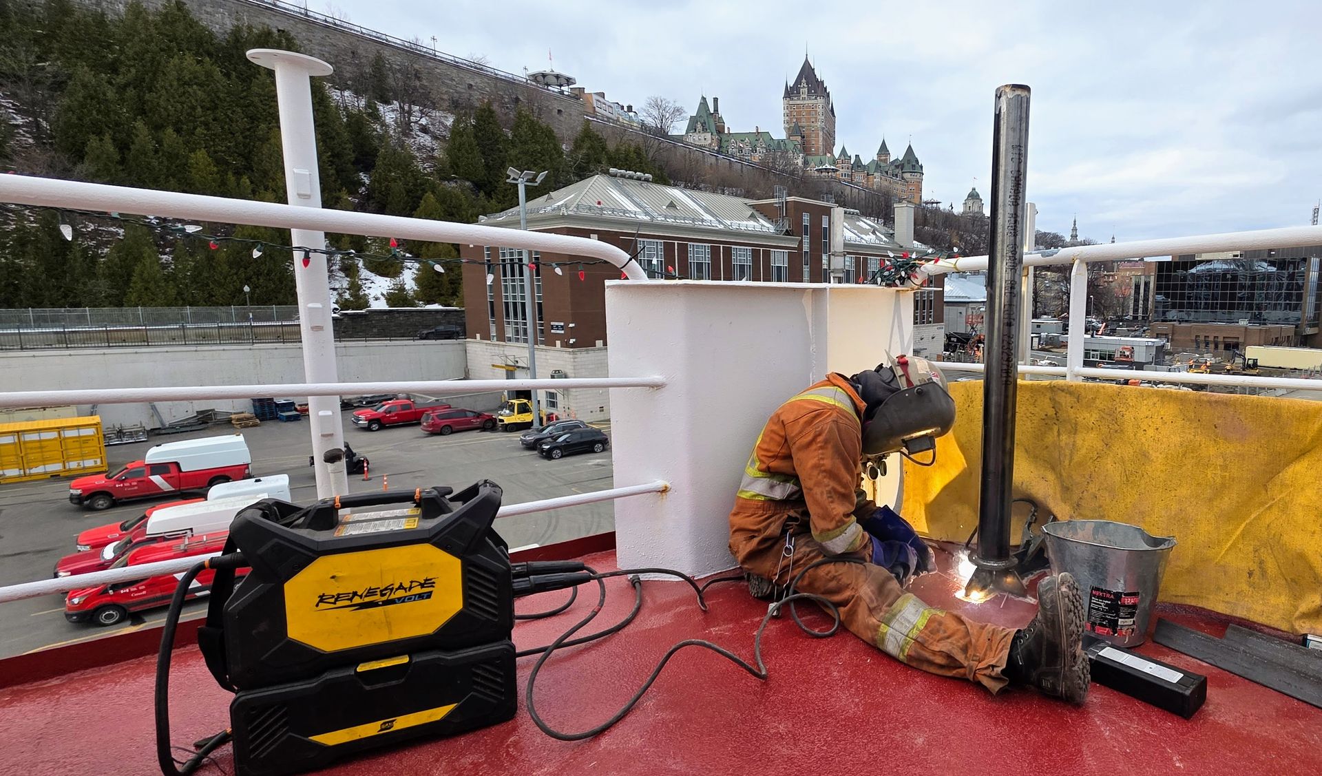 Une personne portant une tenue de soudeur de protection soude un tuyau métallique sur le pont d'un navire, avec le Château Frontenac visible en arrière-plan.