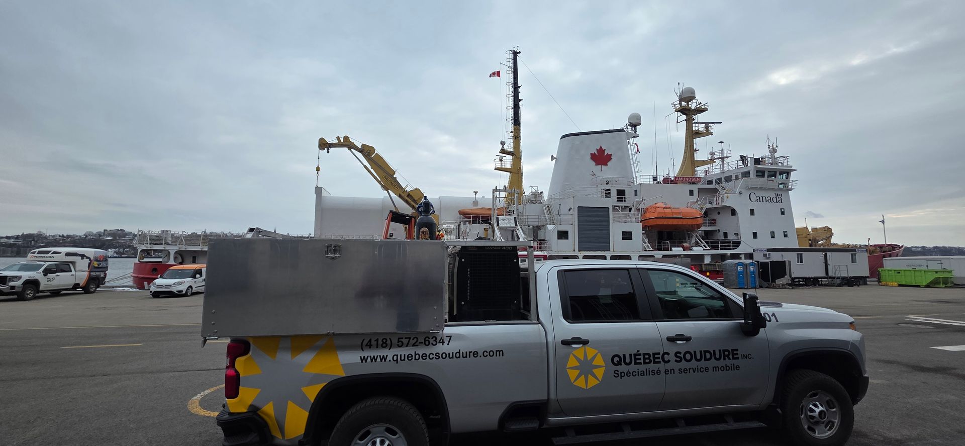 Un camion de service argenté stationné dans un port, avec un grand navire blanc arborant le drapeau canadien en arrière-plan.