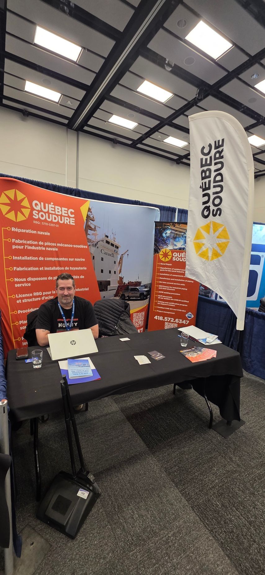 Une personne est assise au stand du salon « Québec Soudure », équipé d'une bannière, d'une table et d'un ordinateur portable, dans un grand hall.