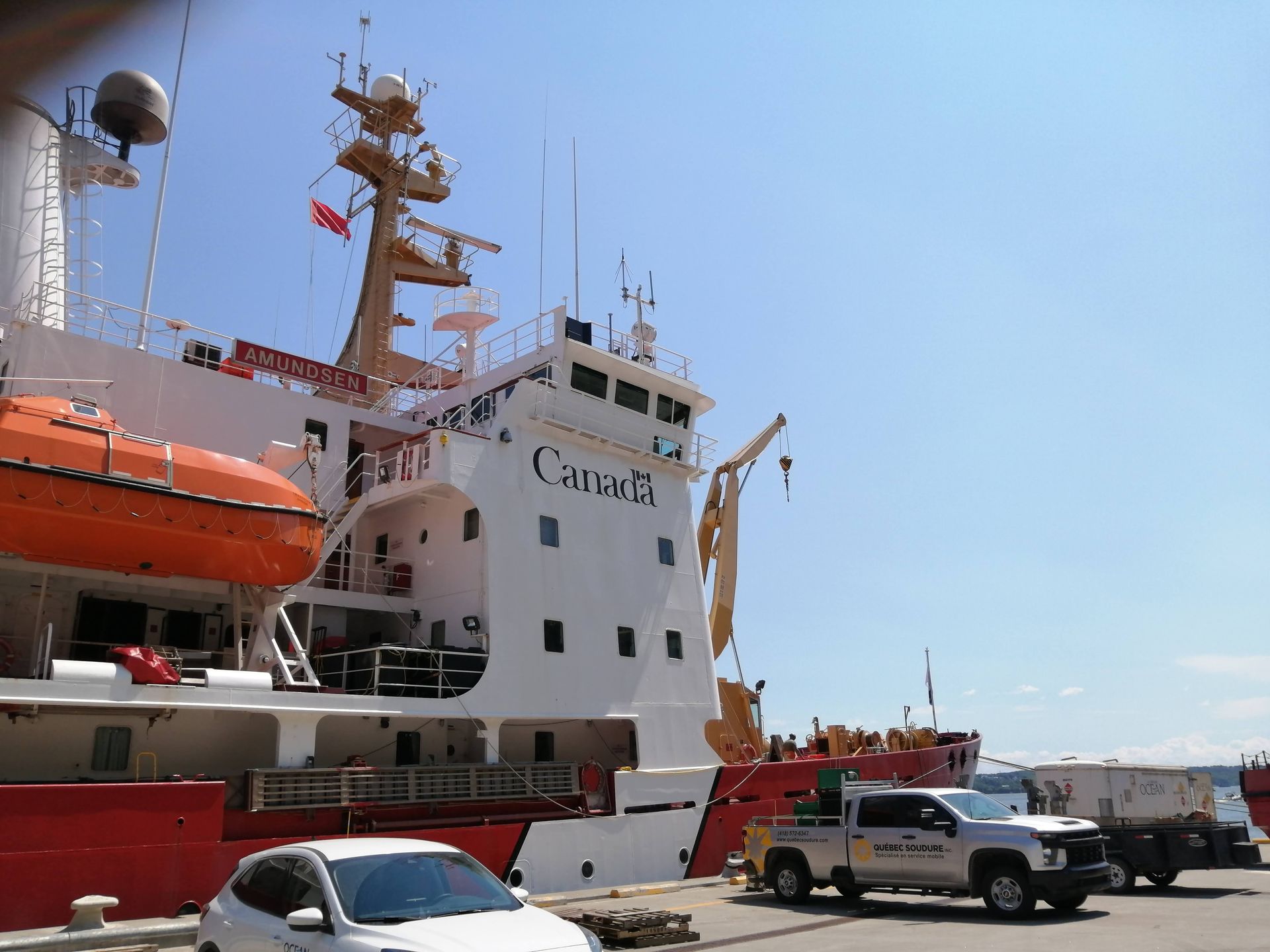 Un navire rouge et blanc des garde-côtes canadiens est amarré à un quai, avec une voiture et un camion garés au premier plan.
