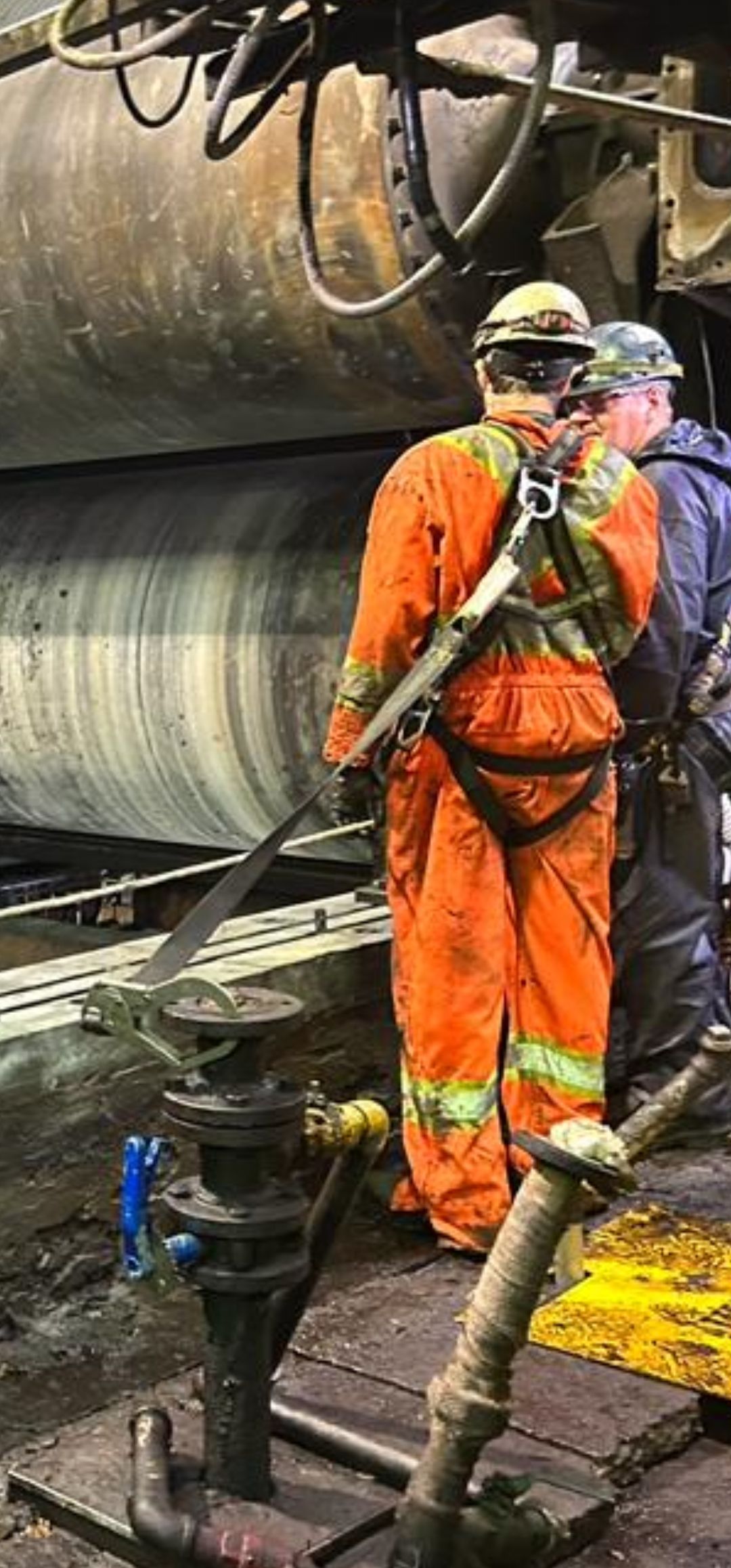 Deux ouvriers, casqués et équipés de protections, se tiennent près de grosses machines dans une usine.