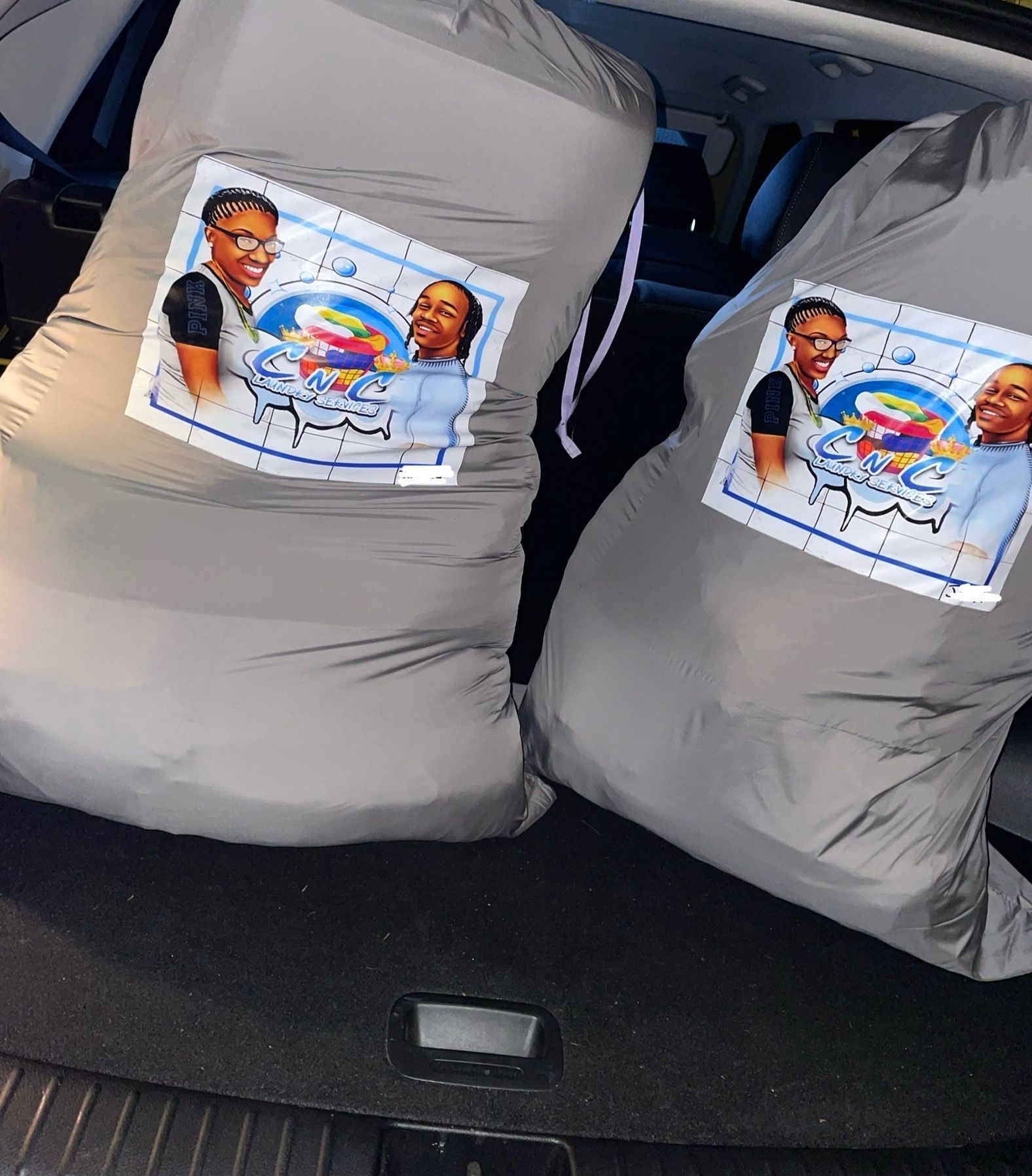 Two large gray laundry bags with a company logo in a car trunk.