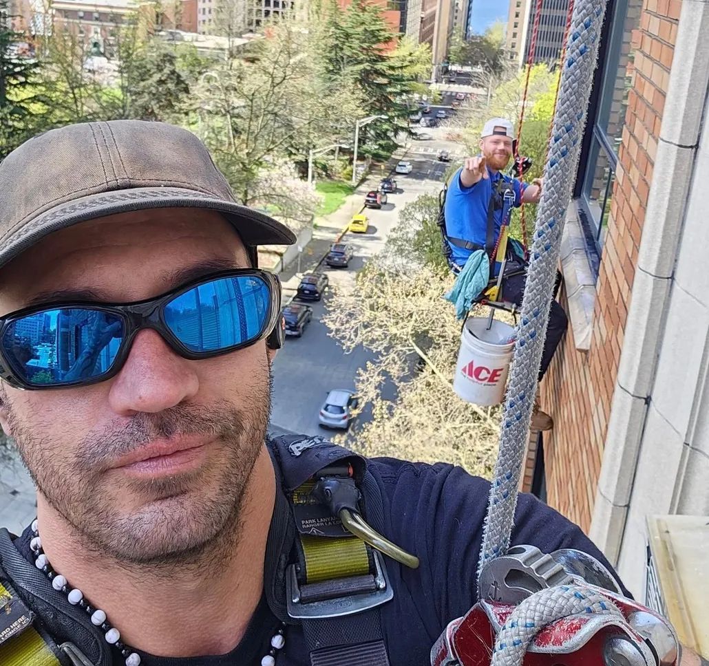 Man in sunglasses selfies while window washing high above city street. Another worker nearby.