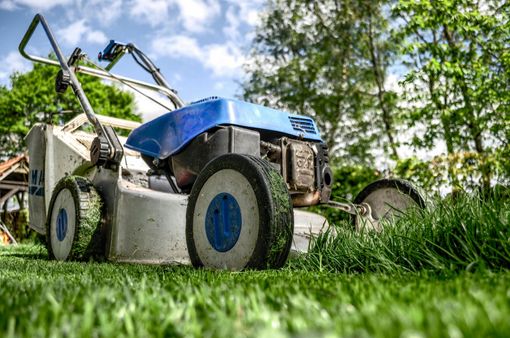 Lawn mower cutting green grass in a yard, blue and white mower, green grass and trees in background.