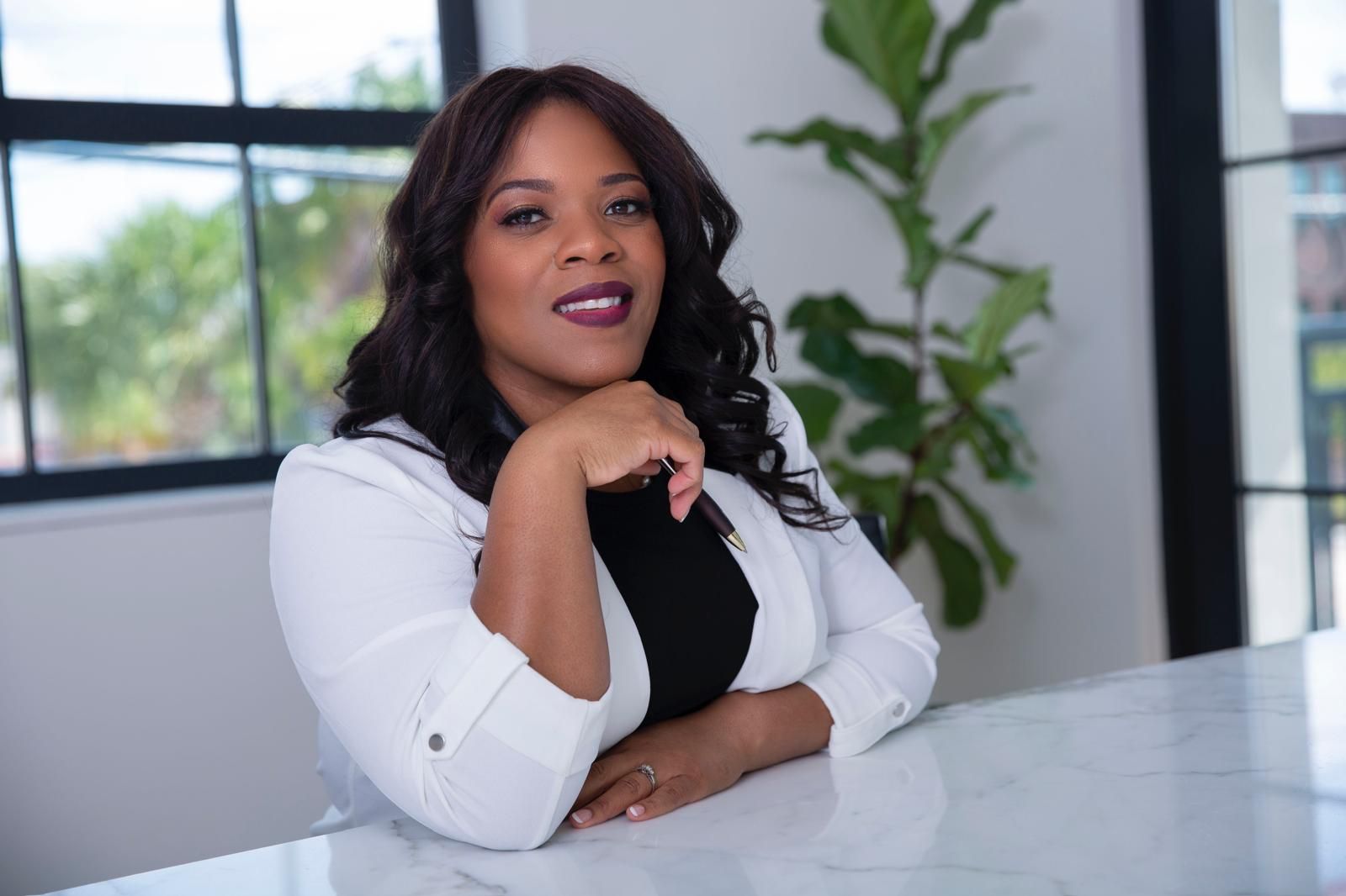 Woman in a white jacket, resting on a white counter; smiling. Interior setting with windows and a plant.