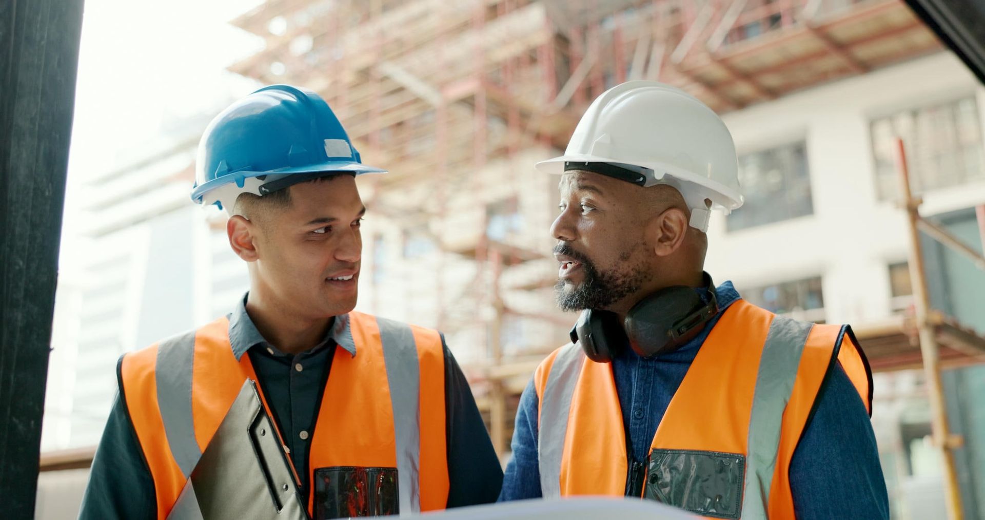 Two construction workers in safety vests and hard hats discussing blueprints on a construction site.