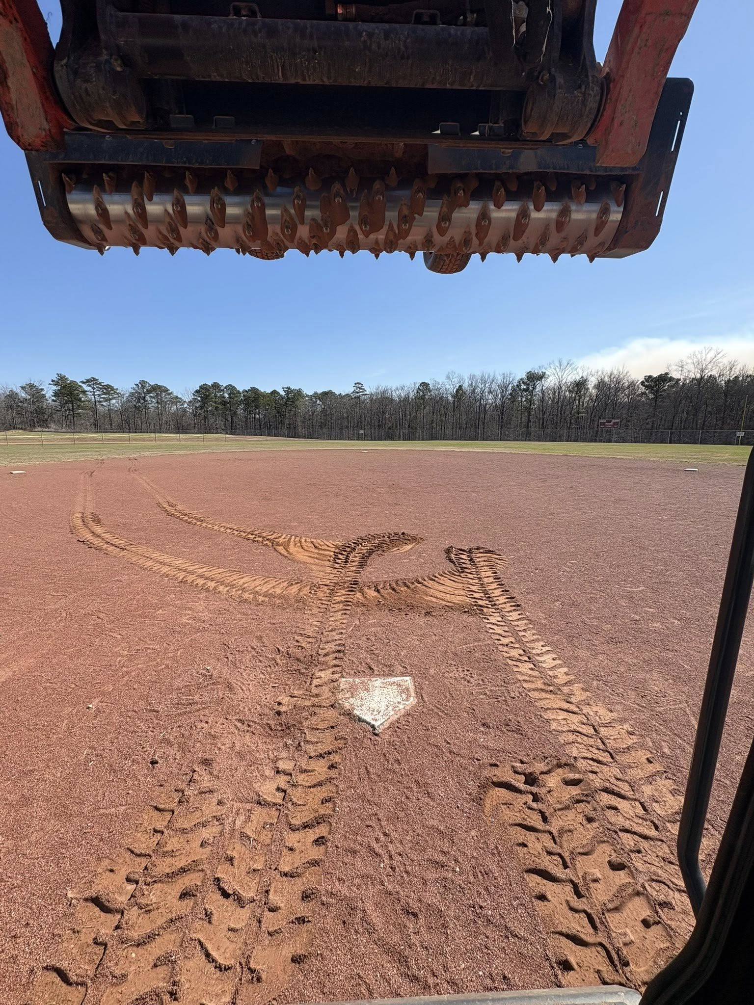 Farming machine above a muddy field with tire tracks and a tree line under a blue sky