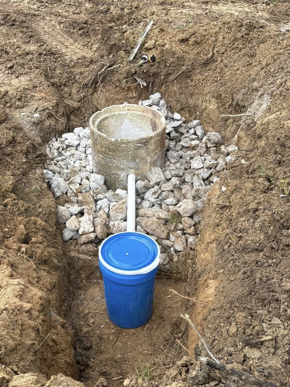 Blue bucket in a dirt trench beside a concrete pipe surrounded by gravel.