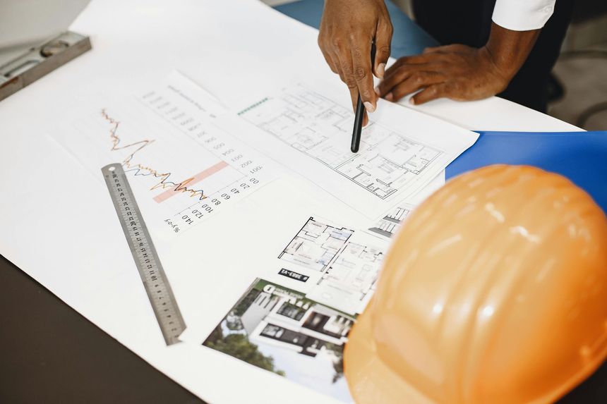 Una persona señala unos planos arquitectónicos sobre un escritorio, junto a un casco naranja y una regla metálica.