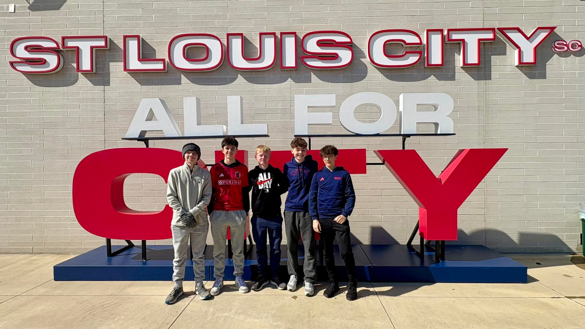 Group of five people posing in front of a "ST LOUIS CITY" sign; the words "ALL FOR CITY" are in front.